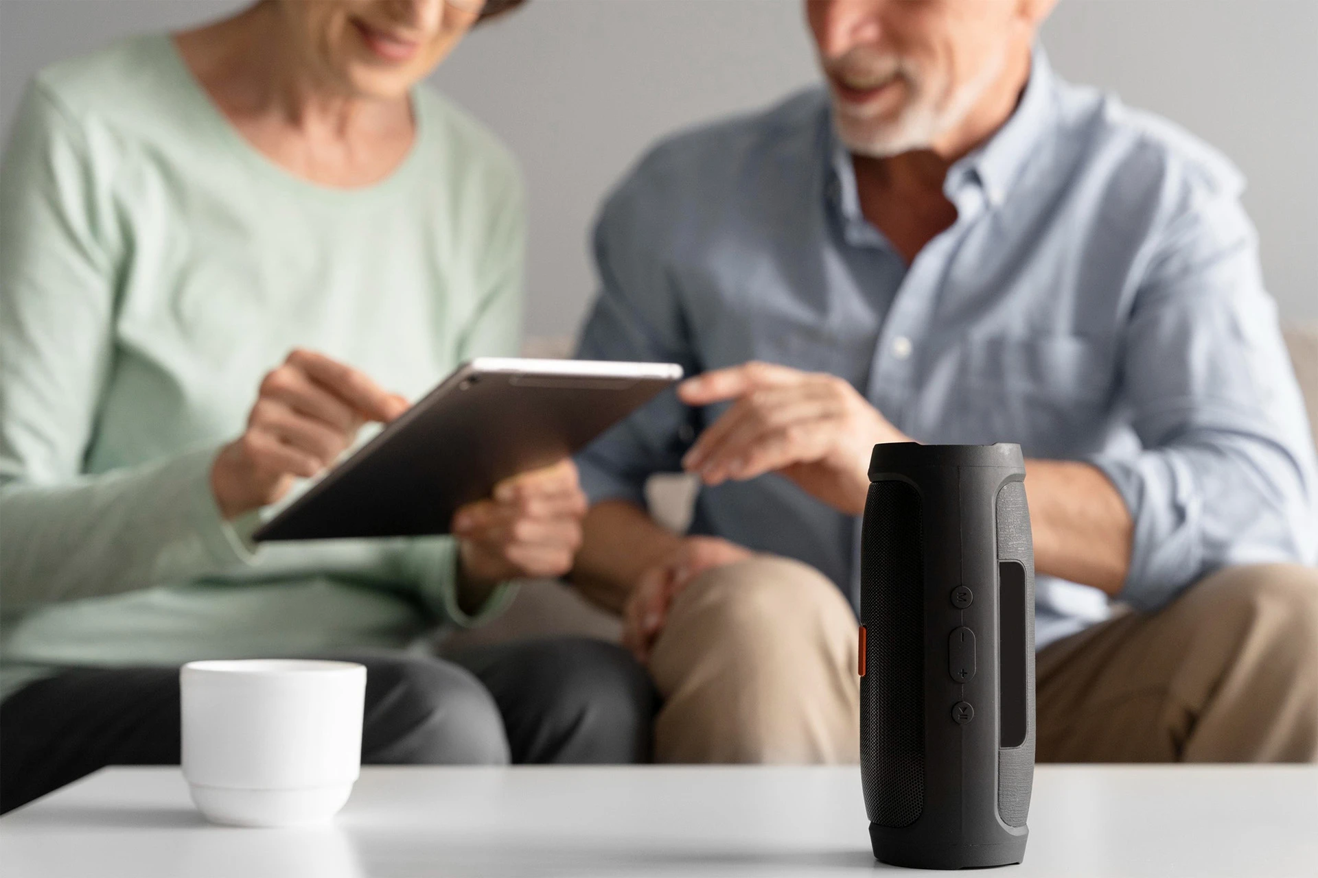 Older couple on a sofa use a tablet, smiling. A Bluetooth speaker and a white mug are on the table. The mood is relaxed and engaged.