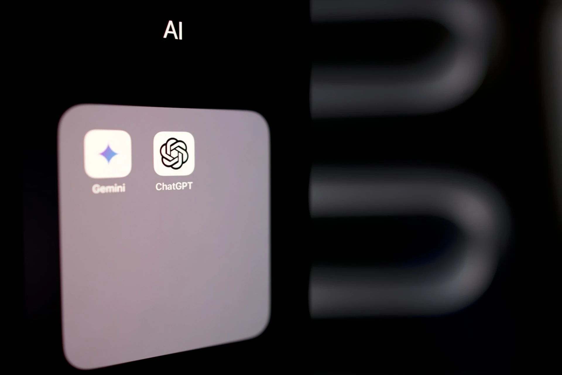 A smartphone displaying a homepage menu labeled “AI,” with two icons for Google Gemini and ChatGPT. Displayed on a dark background.