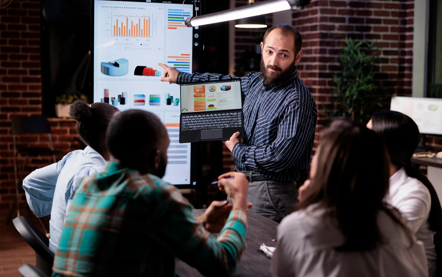 A man presents data to colleagues using a laptop and a large screen showing charts in a modern office.