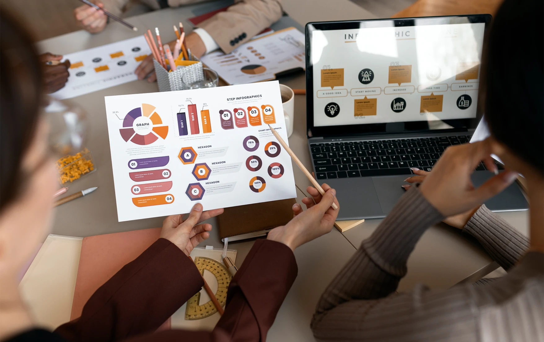 A team discusses data at a table, focusing on print and digital infographics. Laptops and worksheets indicate a collaborative atmosphere.