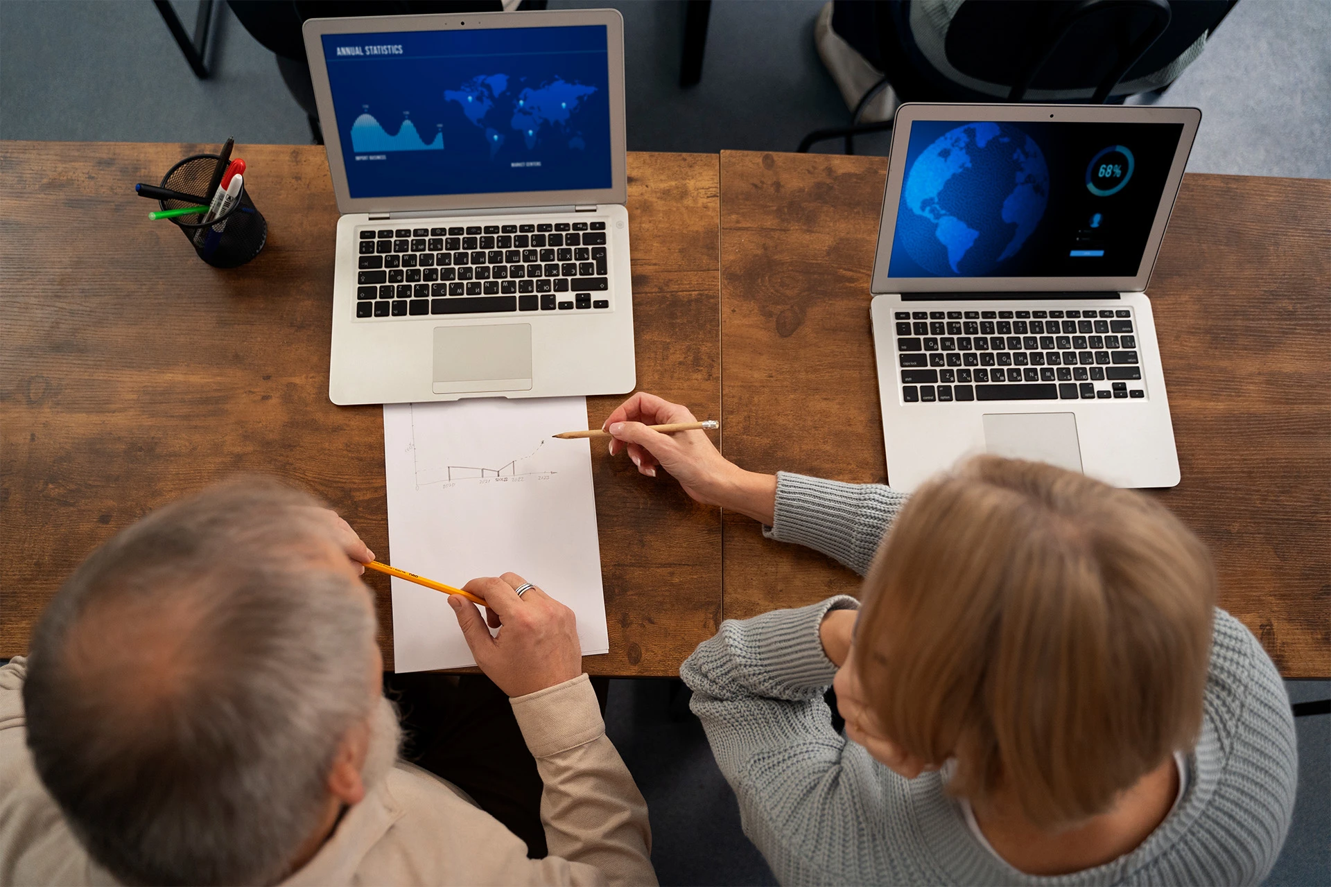 Two people at a wooden table work on laptops showing blue data visuals. They discuss a sketch on paper, showing a collaborative atmosphere.