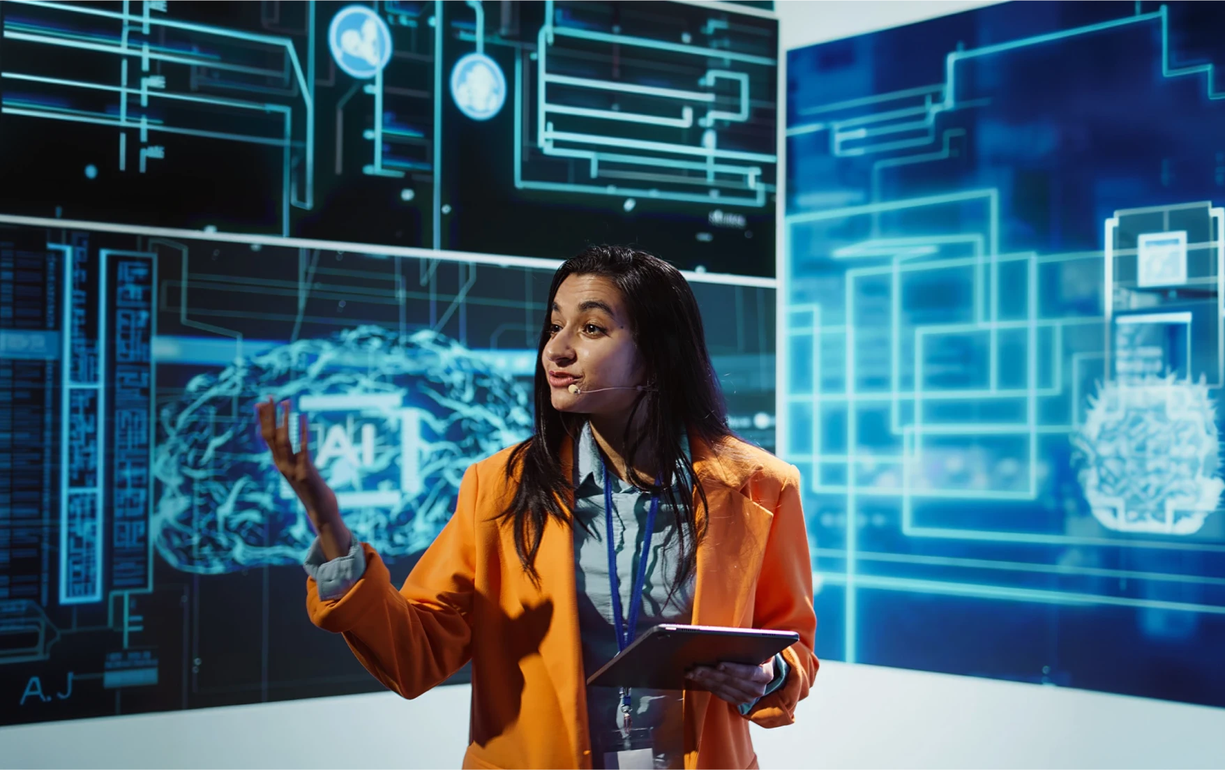 A woman in an orange blazer speaking confidently on stage with a headset and tablet. The background displays digital images of a brain labeled “AI”, indicating a tech presentation.