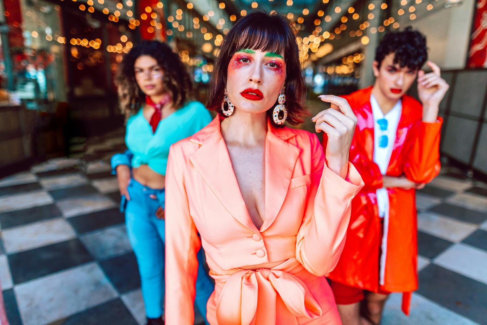 Three stylishly dressed people pose indoors on a black-and-white checkered floor, with warm string lights in the background