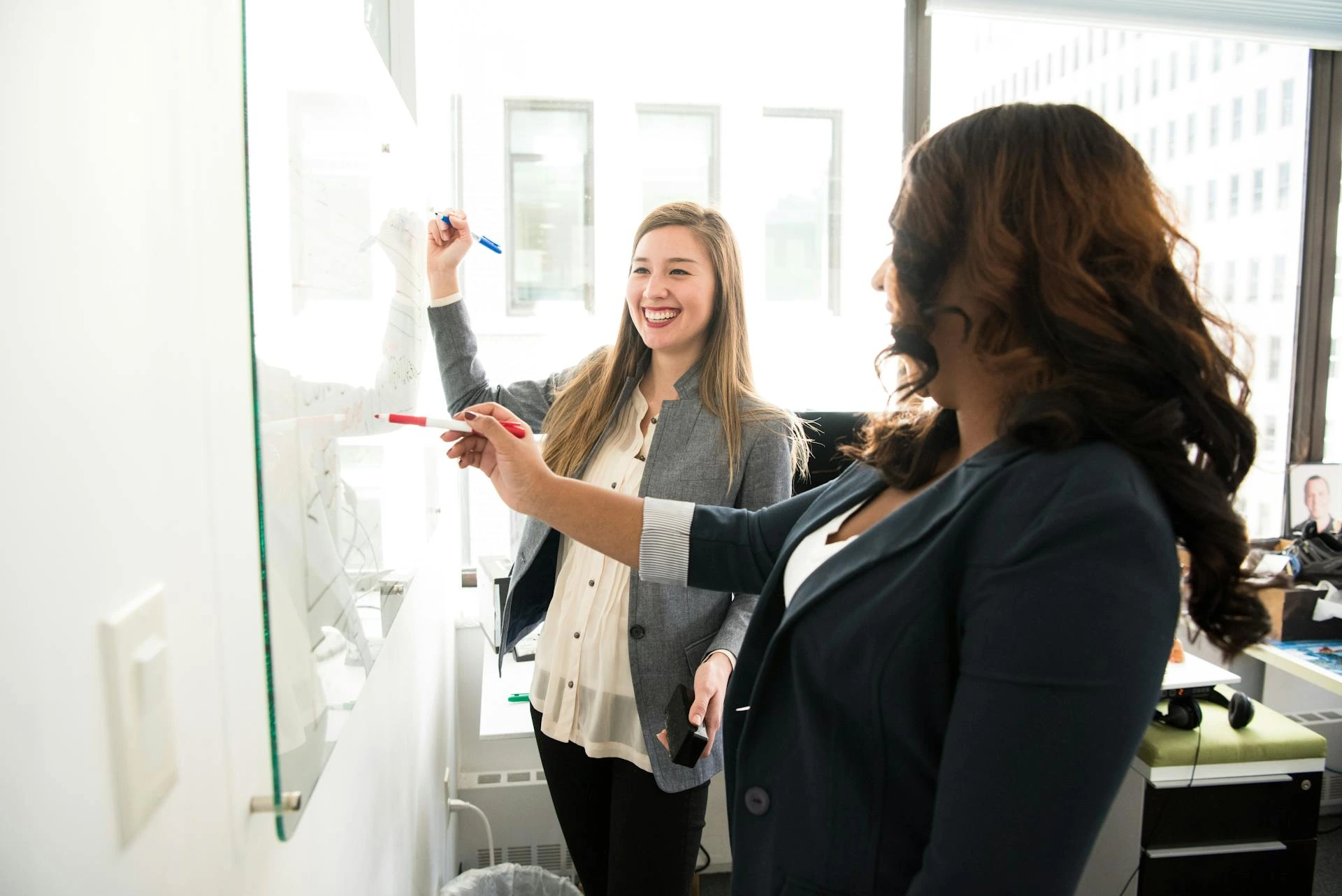 Two women in an office writing on a dry-erase board.