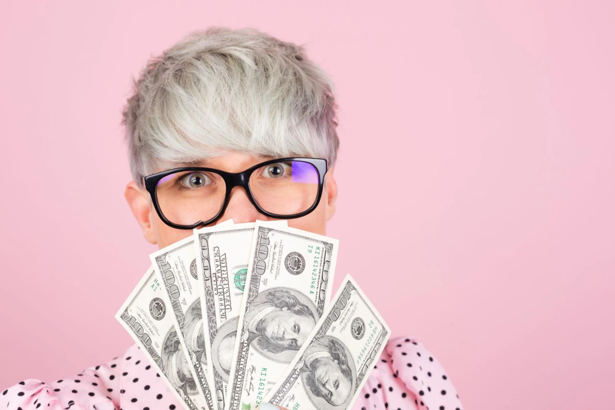 A person with short, textured gray hair is holding a stack of hundred-dollar bills in front of their face. 