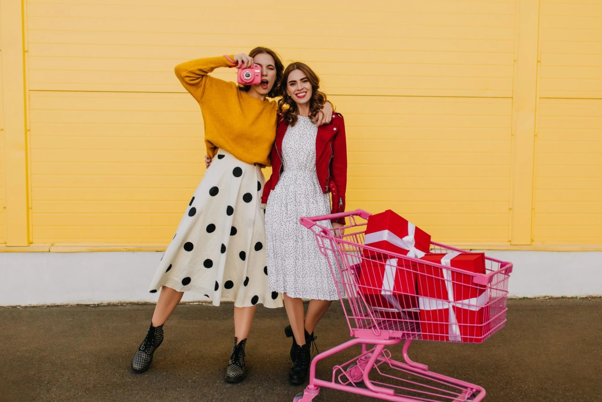 Two young women are seen against a bright yellow backdrop with a pink shopping cart full of wrapped gifts.