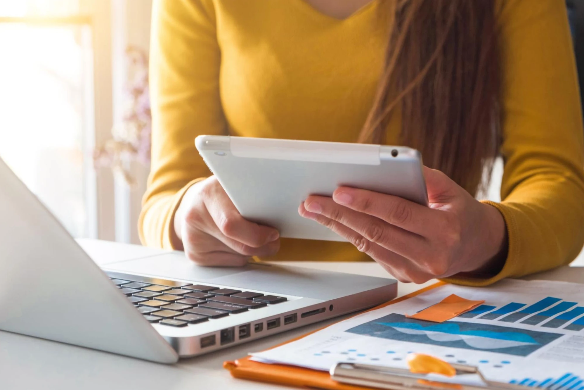 A woman in a yellow shirt holds an iPad at a desk.