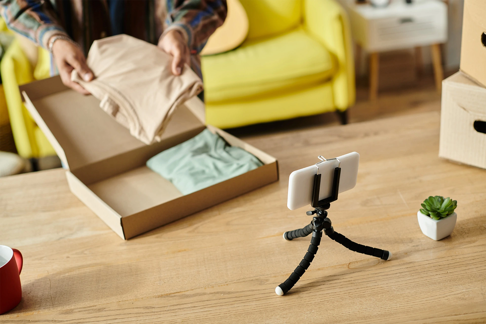 Person taking a pair of shirts out of a cardboard box, filmed by a smartphone on a small tripod. 