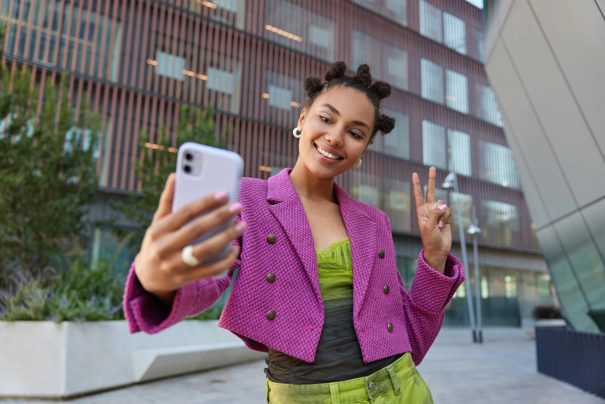 a smiling woman in a vibrant purple jacket takes a selfie outdoors, holding up a peace sign. A lively, contemporary atmosphere.
