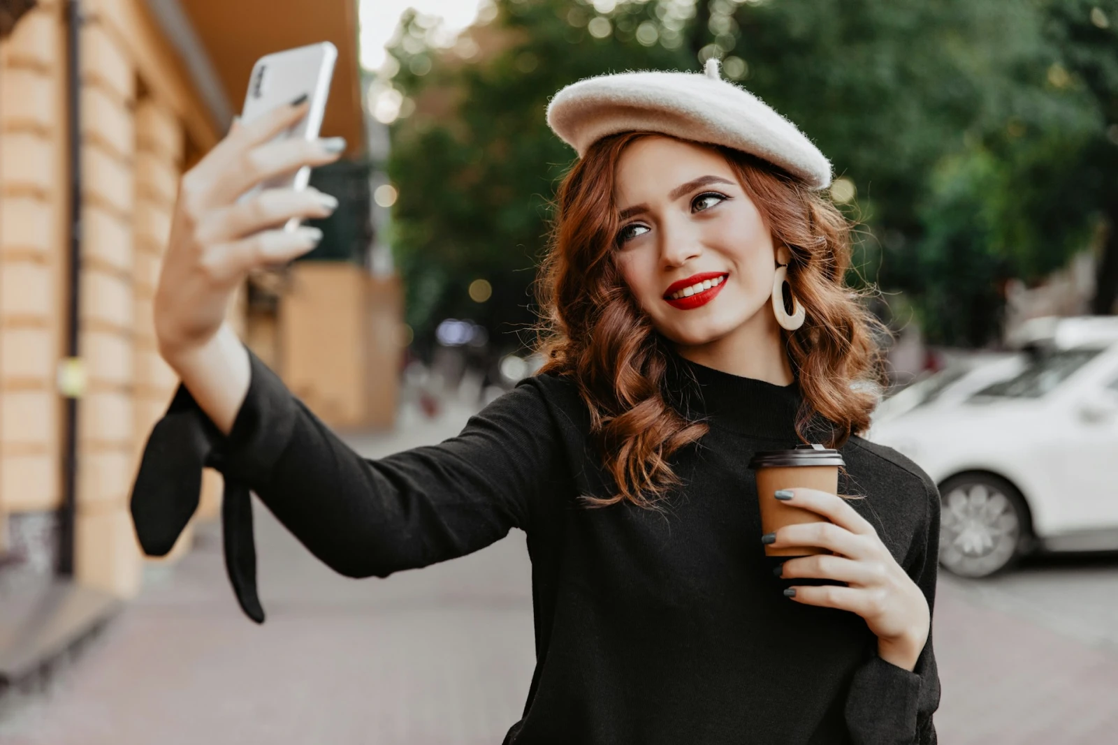 A well-dressed woman taking a selfie while holding coffee on a city street