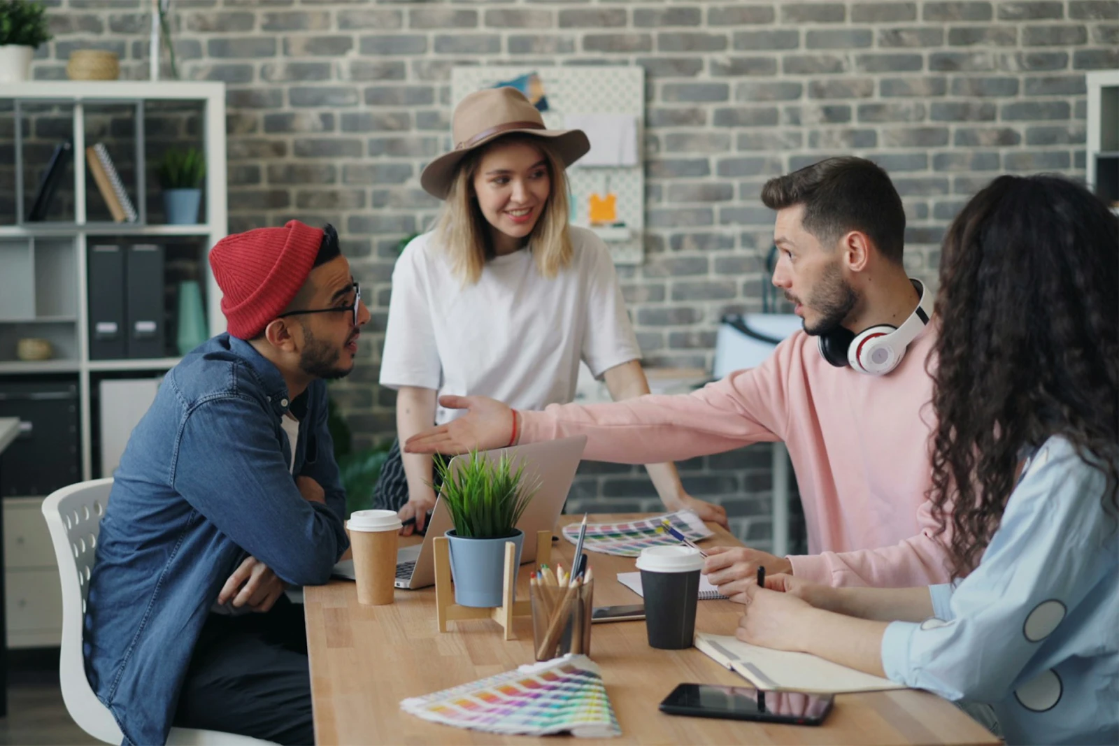 Four people engaging in lively discussion around a wooden table with laptops, color swatches, and coffee cups, set in a modern, creative office