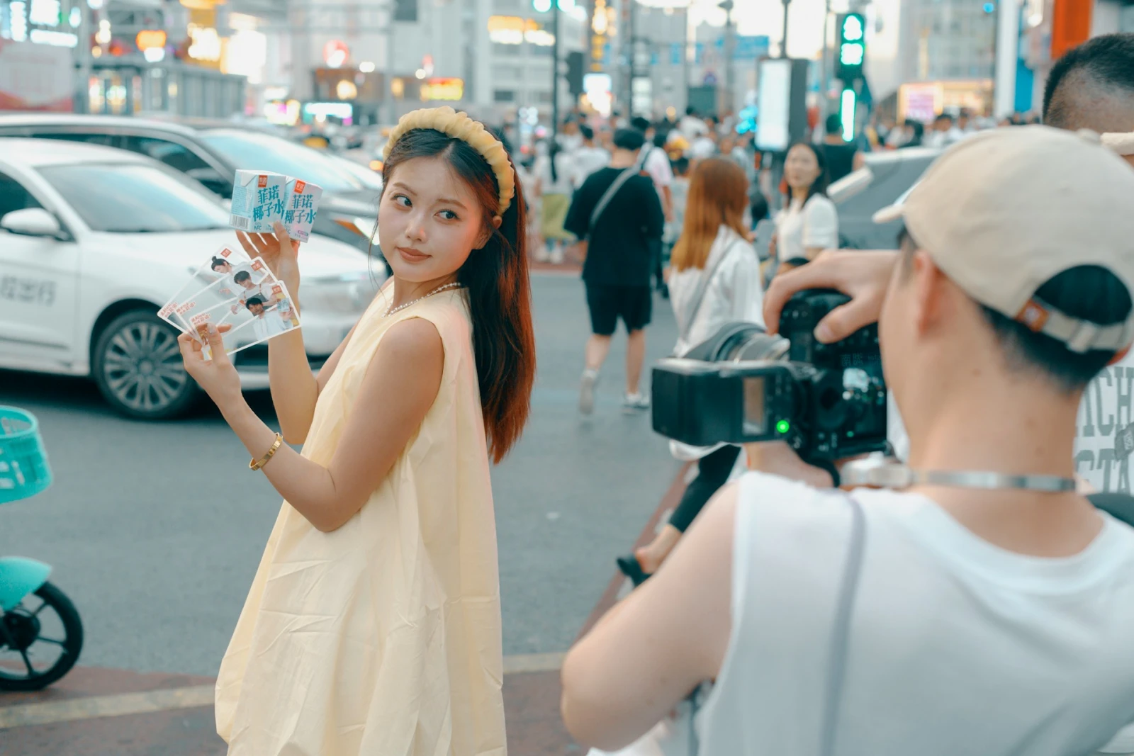 A woman in a yellow dress holds products up to a photographer on a busy city street.
