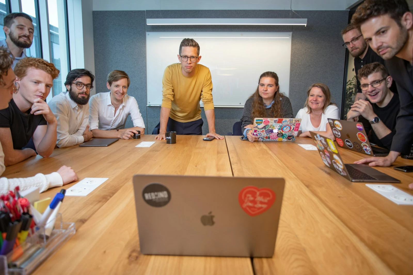 A group of people collaborate in a brightly lit boardroom, all deeply focused on a laptop in the middle of a large wooden table.