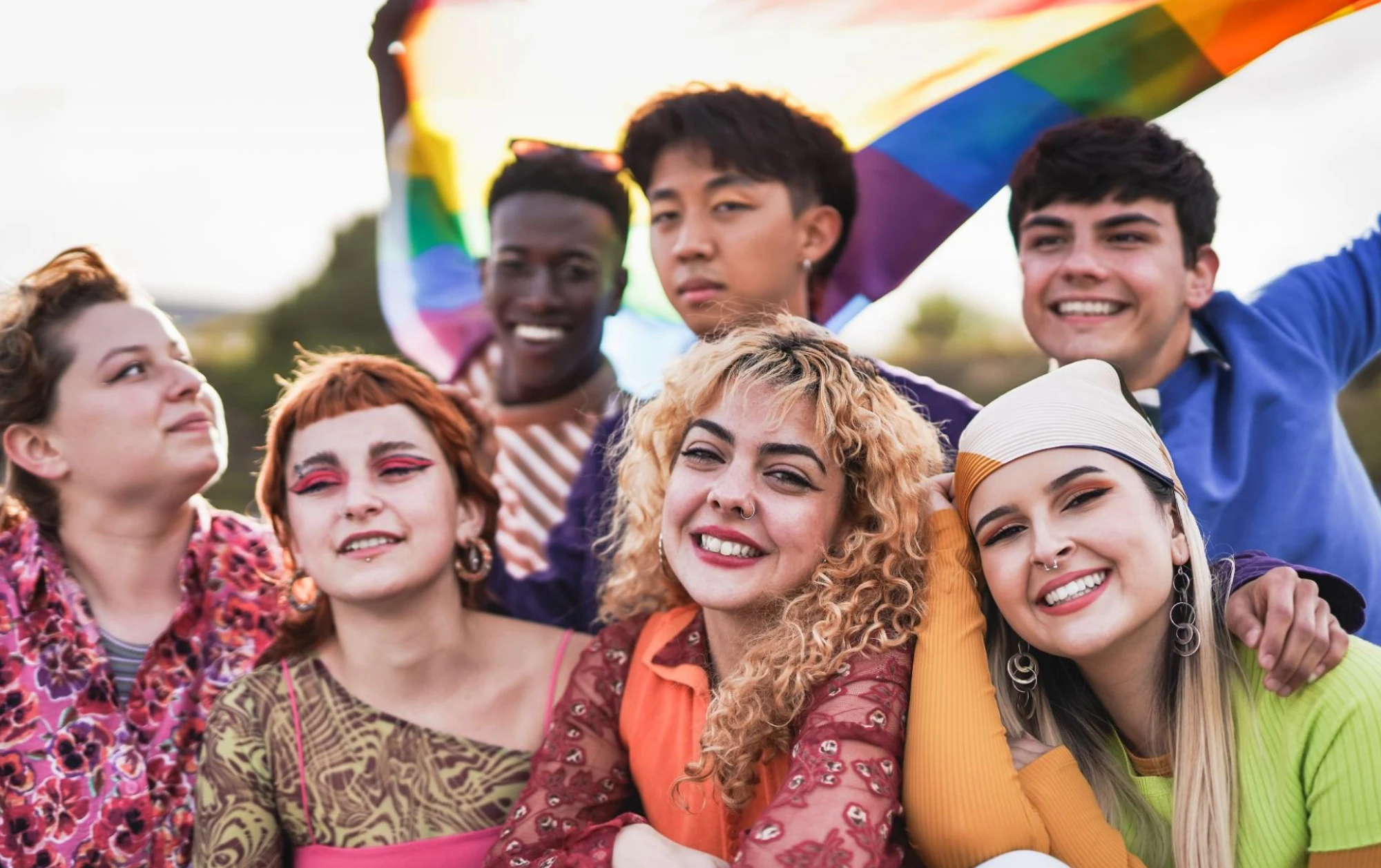 A cheerful group of six young people smiles for the camera, one holding a rainbow flag.