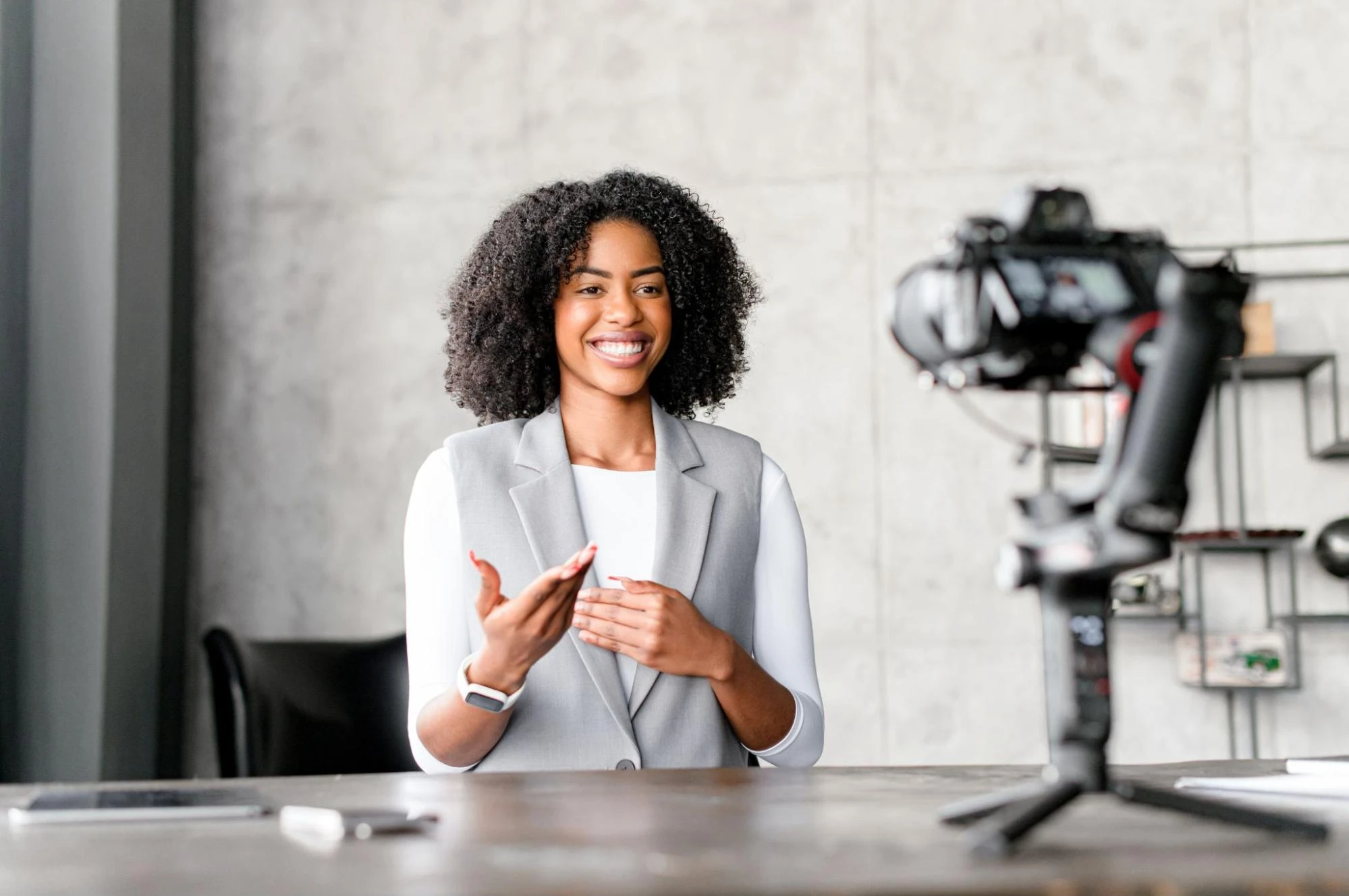 A young woman in a grey vest speaks to a camera sitting on the desk.