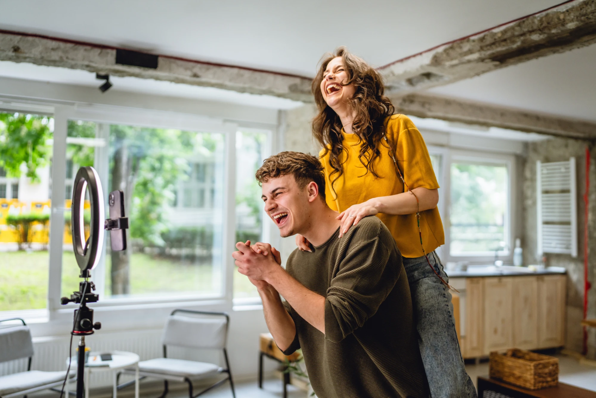 Two young adults laughing together while filming content in a bright modern room, standing beside a ring light and smartphone setup