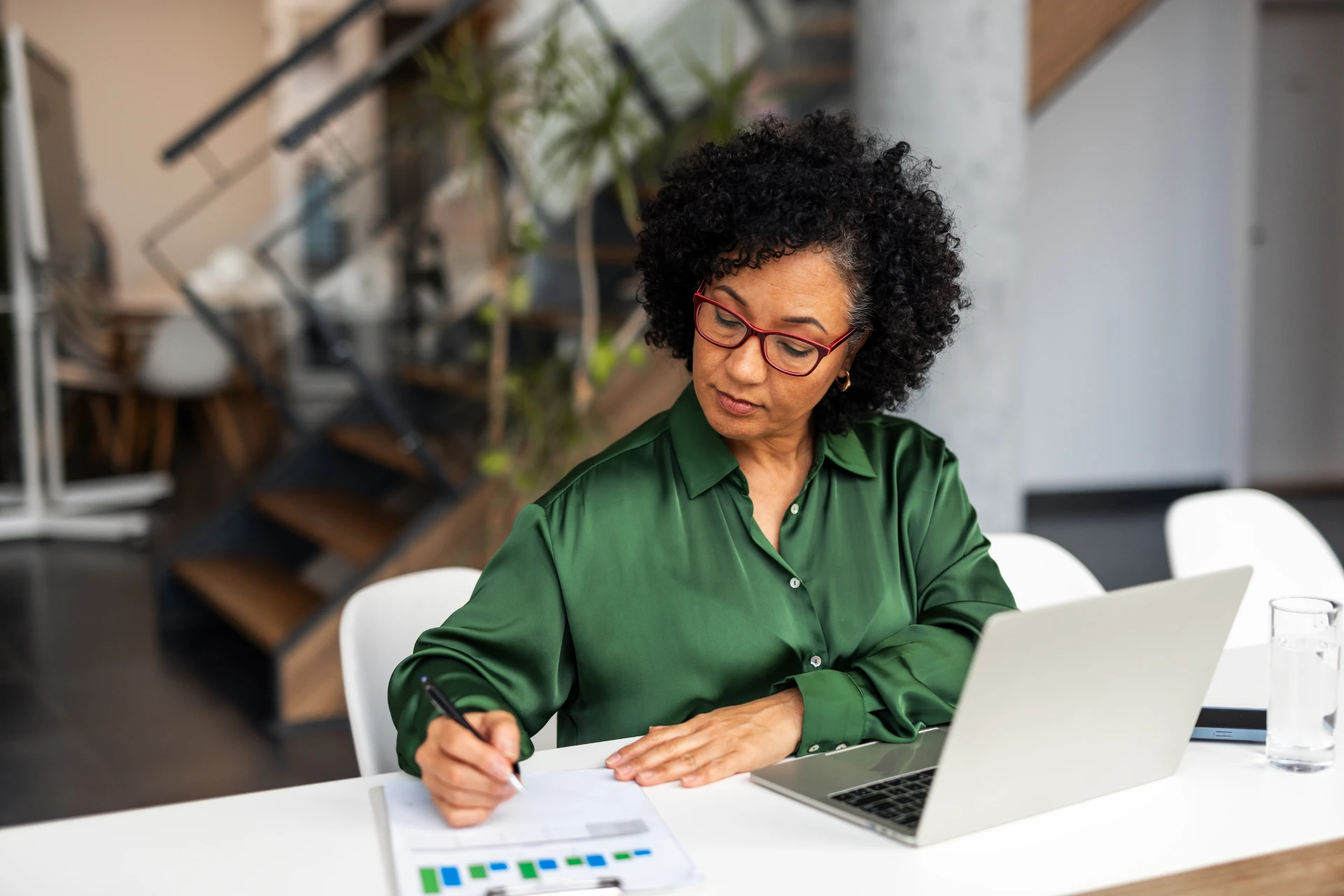 Woman in green blouse and red glasses writing on a document beside a laptop in a modern office