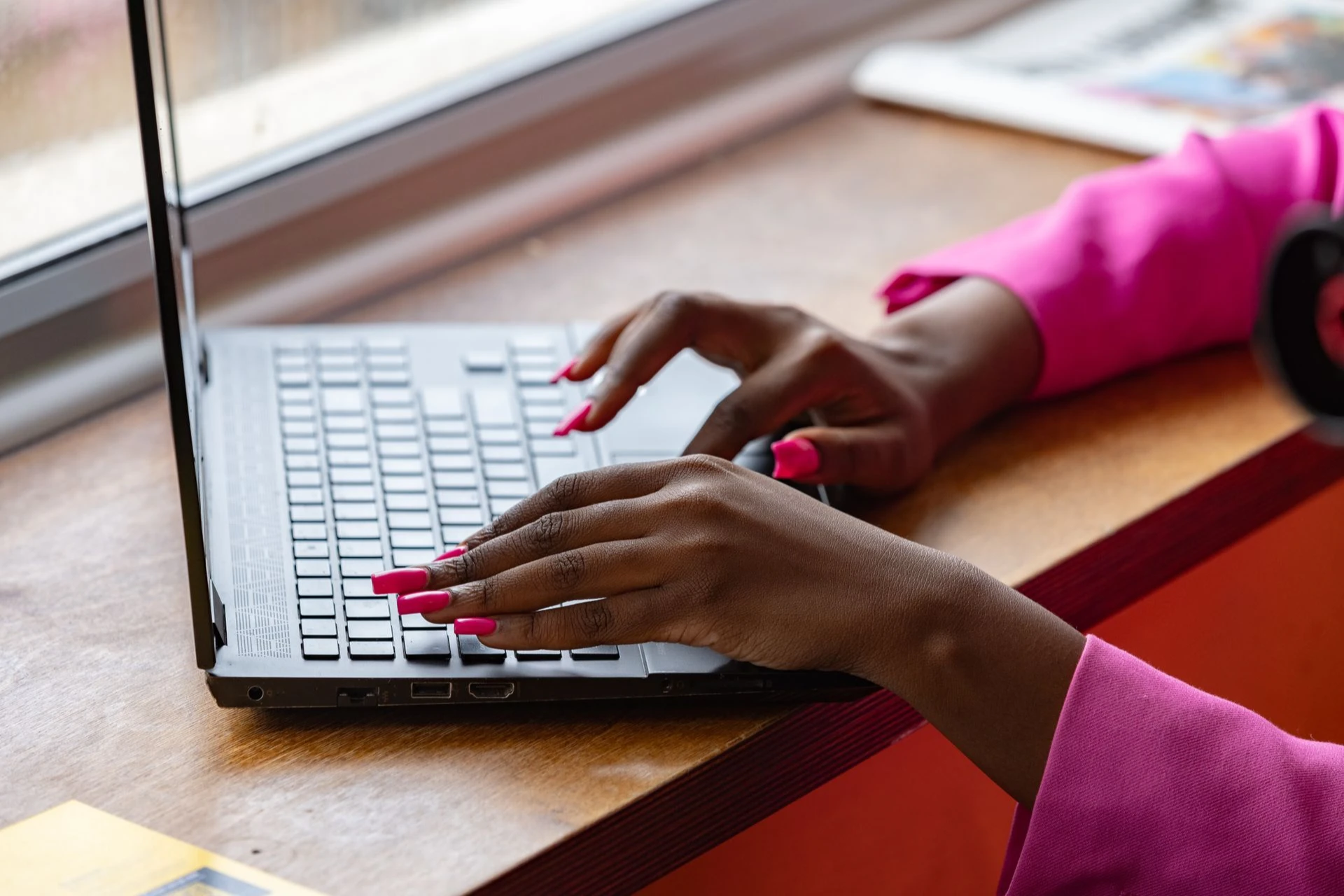 A woman with vibrant pink nails types on a laptop near a window, wearing a bright pink sleeve