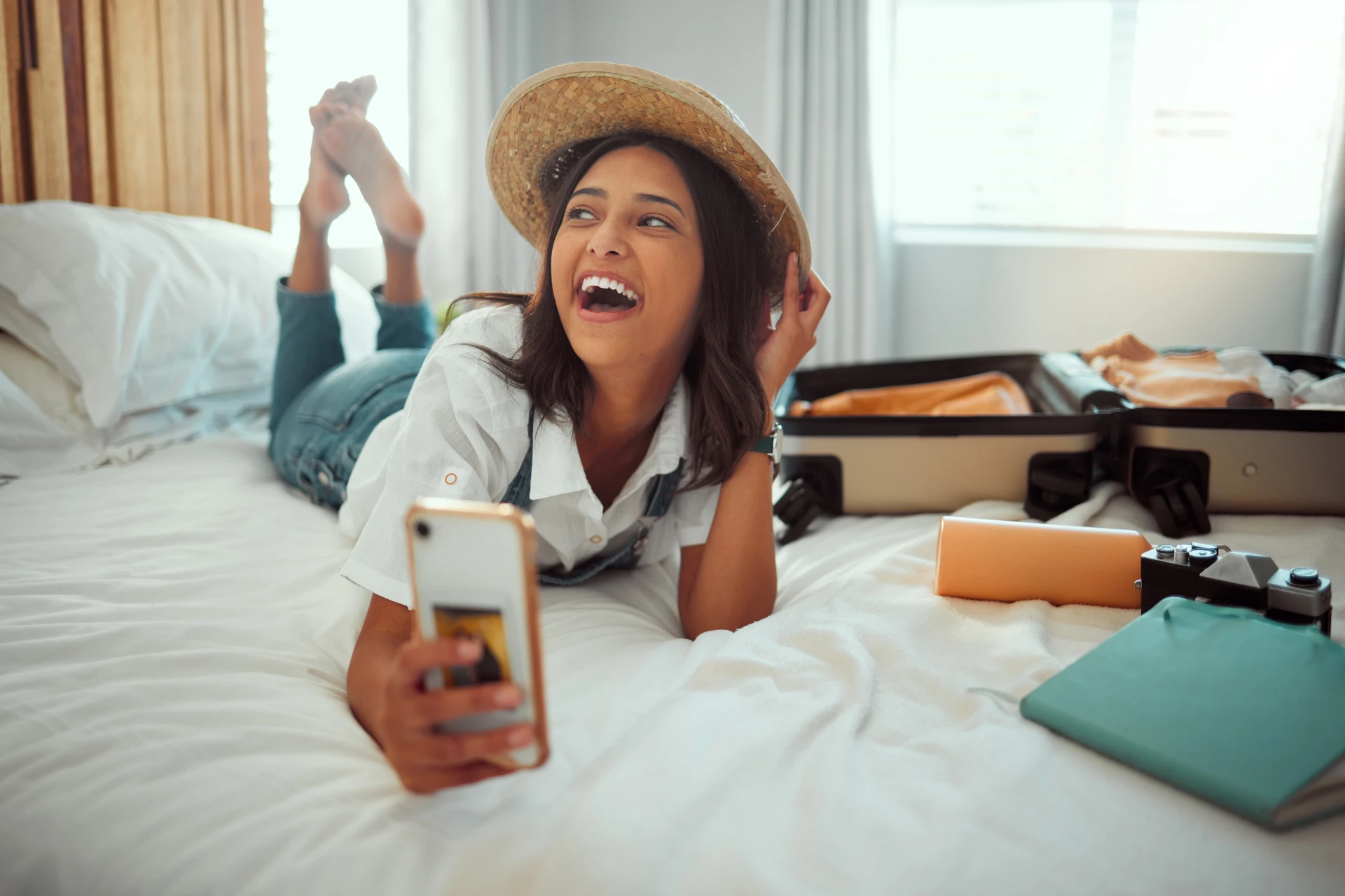 A young woman with dark wavy hair lying on a white bed and smiles while holding a smartphone