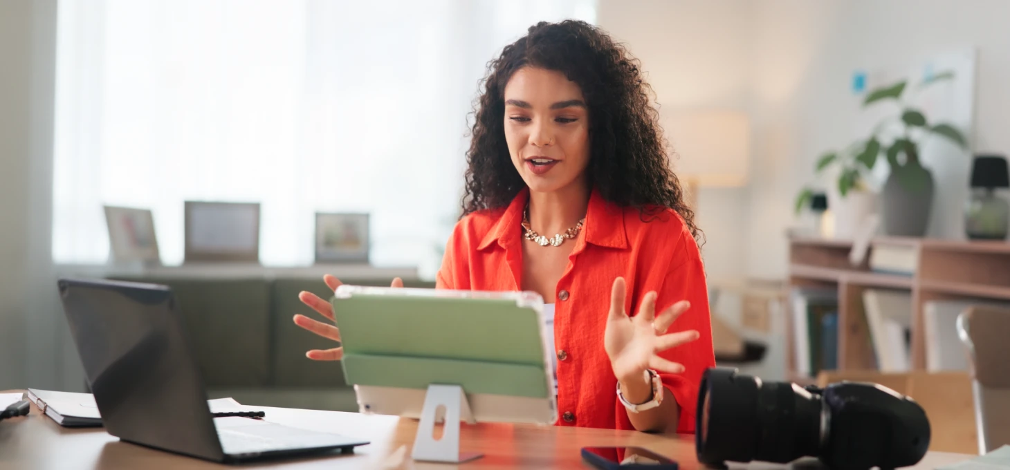 Woman in an orange shirt speaking to a tablet camera while recording content at her desk