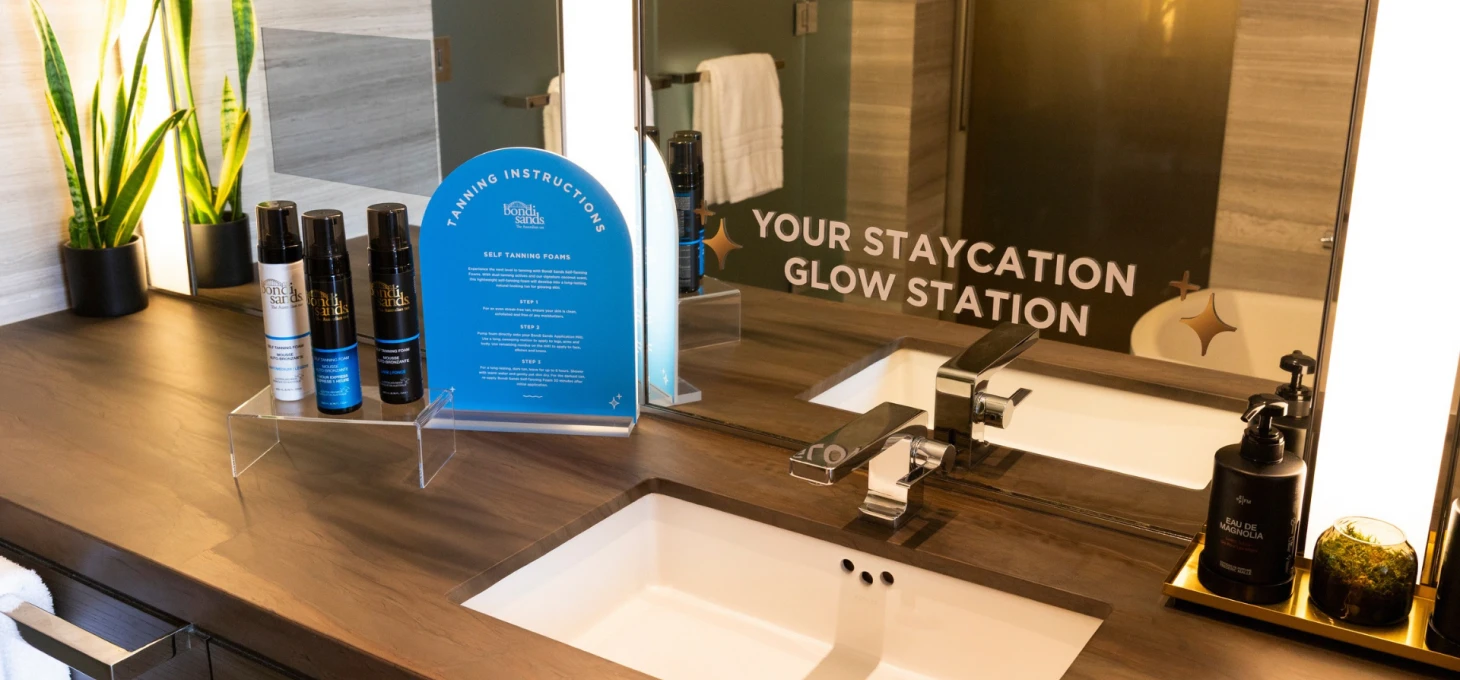 A modern hotel bathroom vanity with a white rectangular sink, chrome faucet, wood countertop, and large mirror displaying the text “YOUR STAYCATION GLOW STATION.”