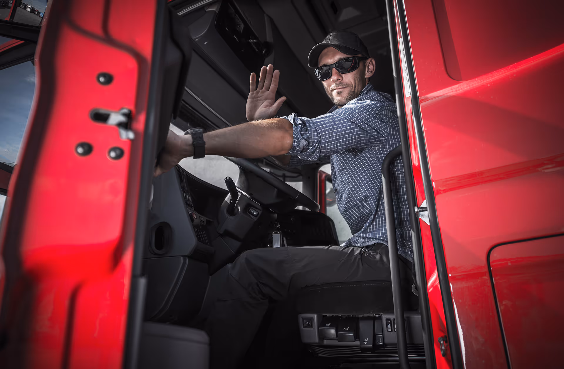 Truck driver in sunglasses sitting in red semi-truck cab, waving