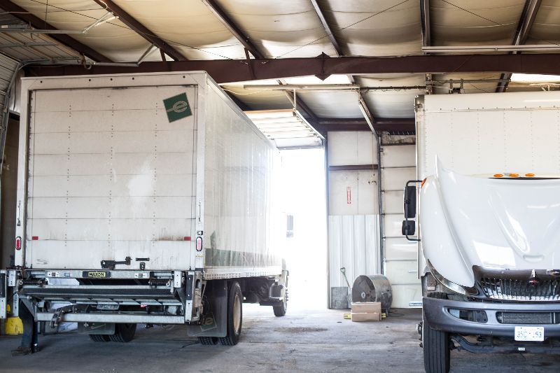 Two box trucks inside a repair shop, one with hood open and another backed in near open bay door, under metal ceiling.