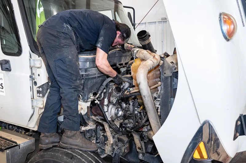Fleet maintenance technician repairs a heavy-duty truck engine with the hood open in a service shop.