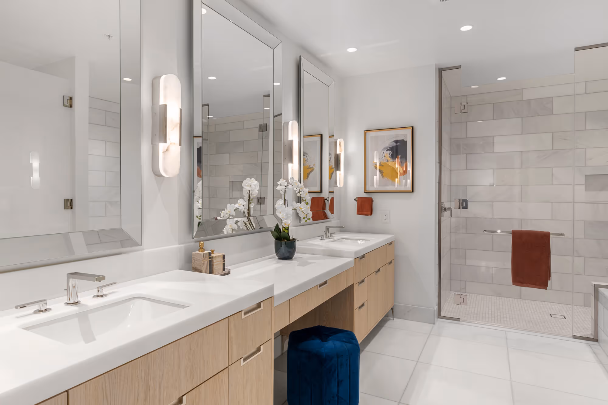 Residence bathroom with double sinks, large mirrors, light wood cabinets, glass-enclosed shower, and blue stool.