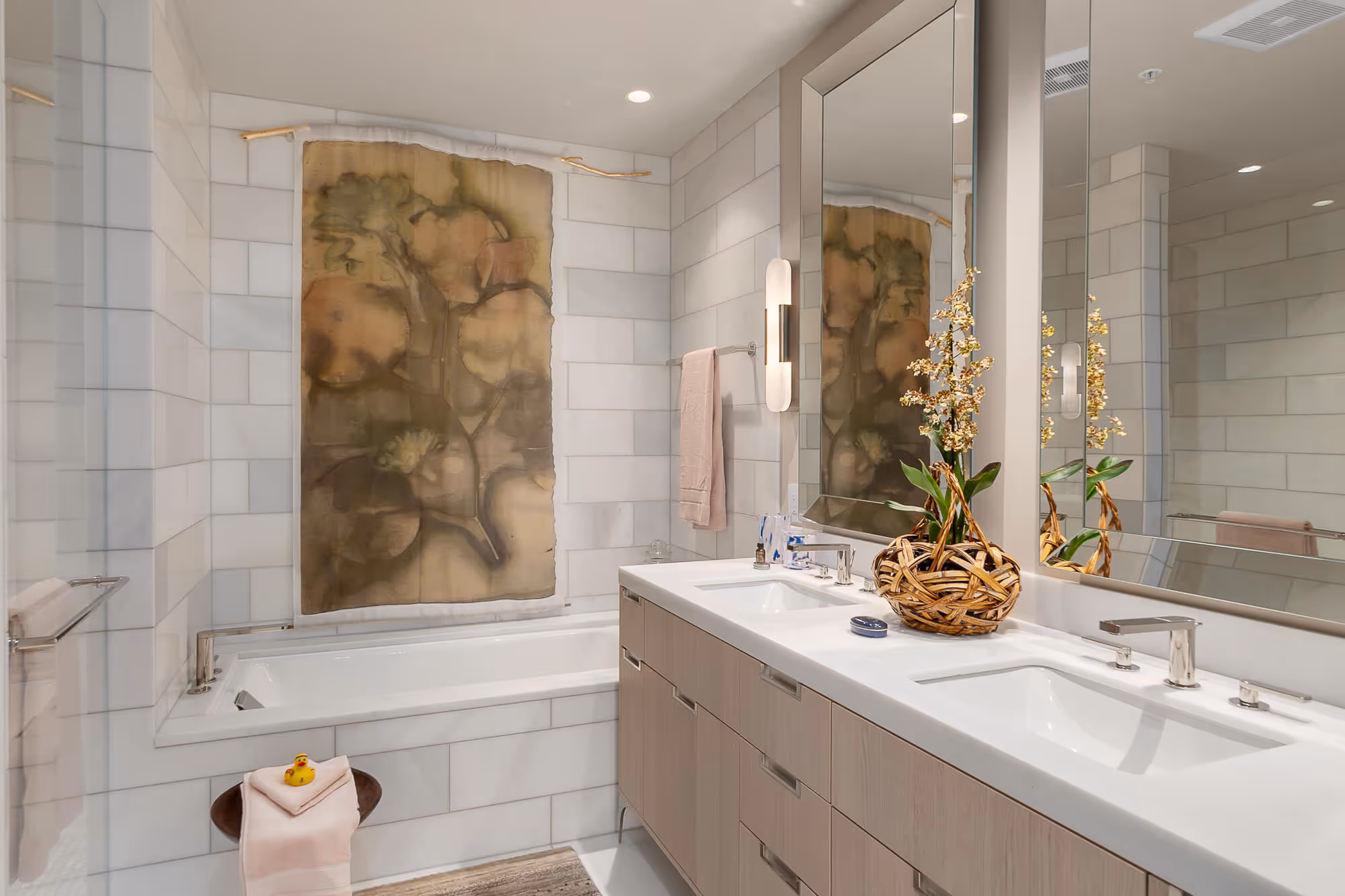 Residence bathroom with white tiled walls, a large white bathtub, double sink vanity with light wood cabinets, and a decorative floral arrangement in a woven basket.