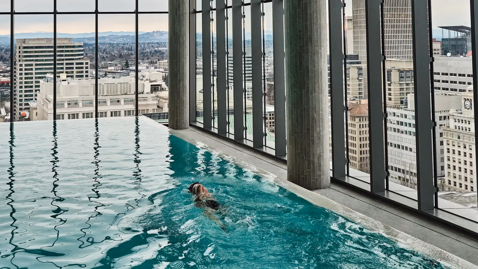 Person swimming in Hidden Mountain Lake infinity pool overlooking a cityscape through large floor-to-ceiling windows.