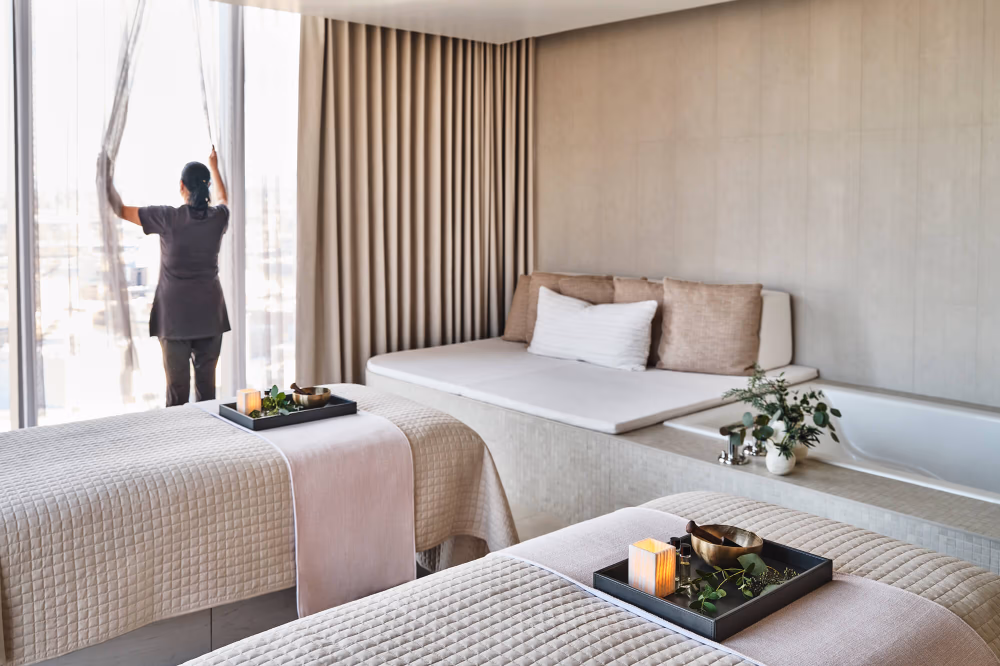 Room at The Ritz-Carlton Spa, Portland with two massage beds, decorative trays with candles and a person opening sheer curtains to a window.