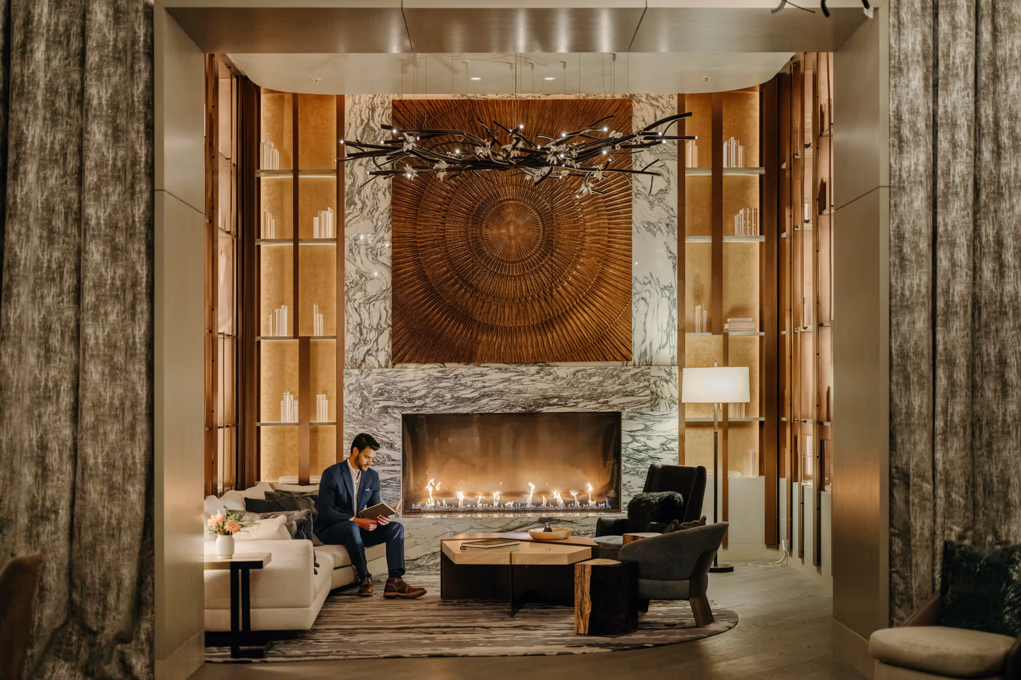 Man in a navy suit sitting on a cream sofa reading a book beside a modern fireplace in the stylish Ritz-Carlton, Portland hotel lobby library with wooden and marble decor.