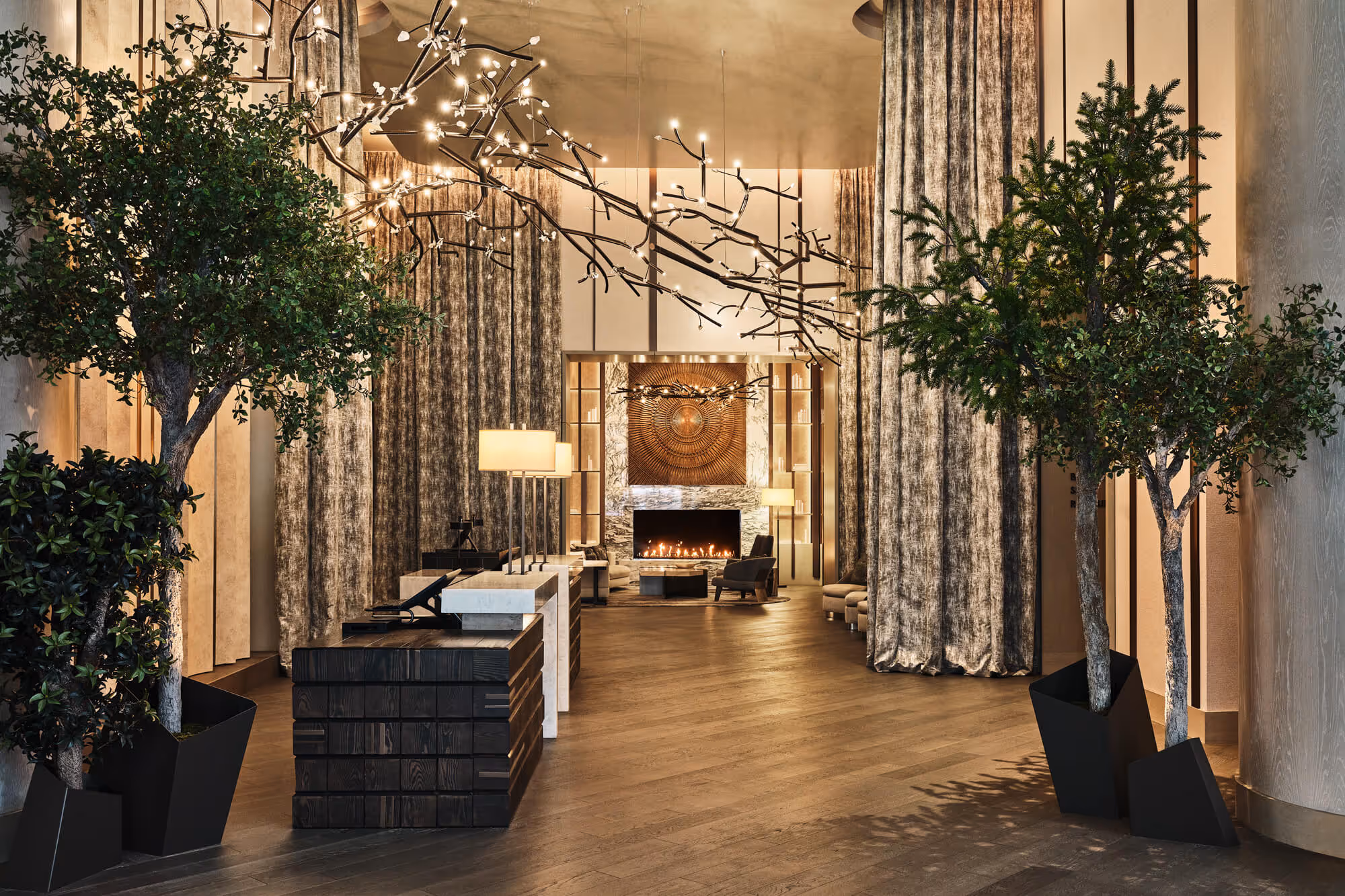 Elegant The Ritz-Carlton, Portland hotel lobby with modern black and white reception desks, large potted trees, hanging branch lighting, tall curtains, and a cozy lit fireplace framed by a decorative wooden panel.
