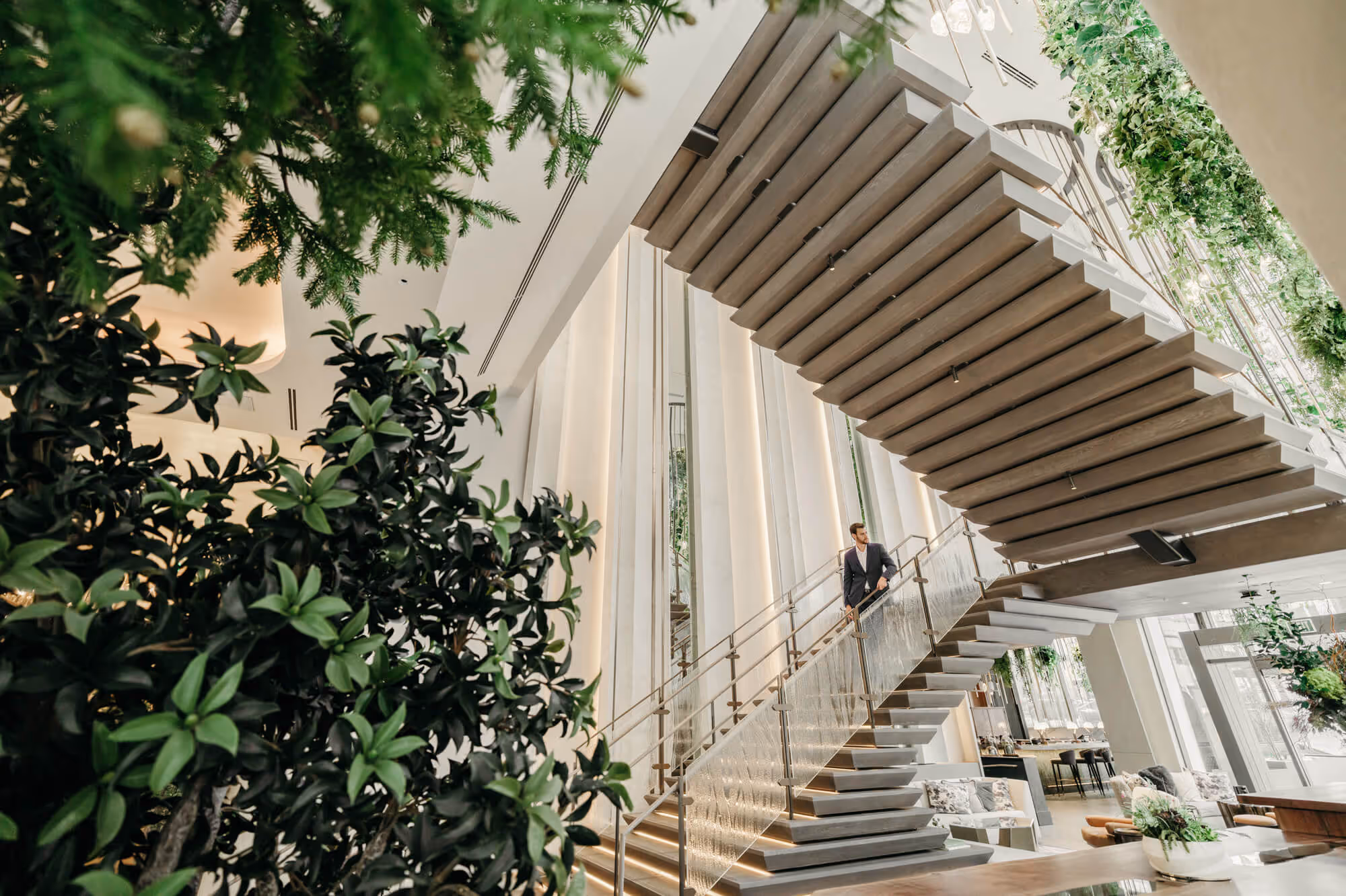 Dramatic staircase in the Grand Staircase Lounge at The Ritz-Carlton, Portland hotel with glass railing surrounded by lush green plants and a man in a suit descending the stairs.