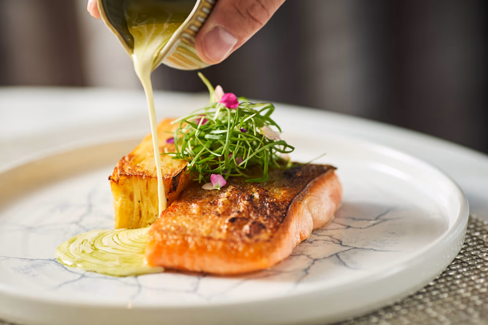 Hand pouring creamy sauce onto a plate with a seared salmon fillet topped with greens and a roasted potato wedge at Bellpine
