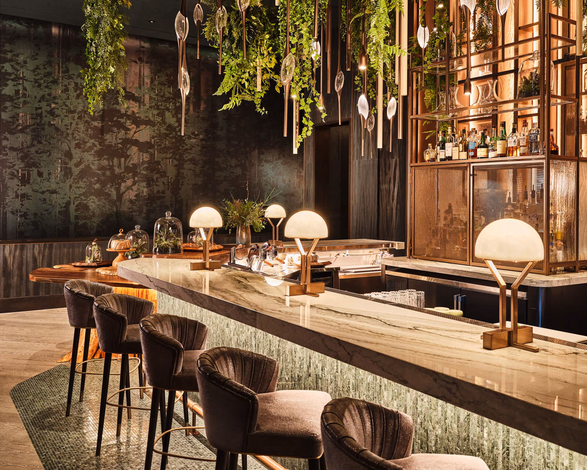 Modern bar interior with a marble countertop, stylish cushioned bar stools, hanging plants, and warm globe lamps at Meadowrue