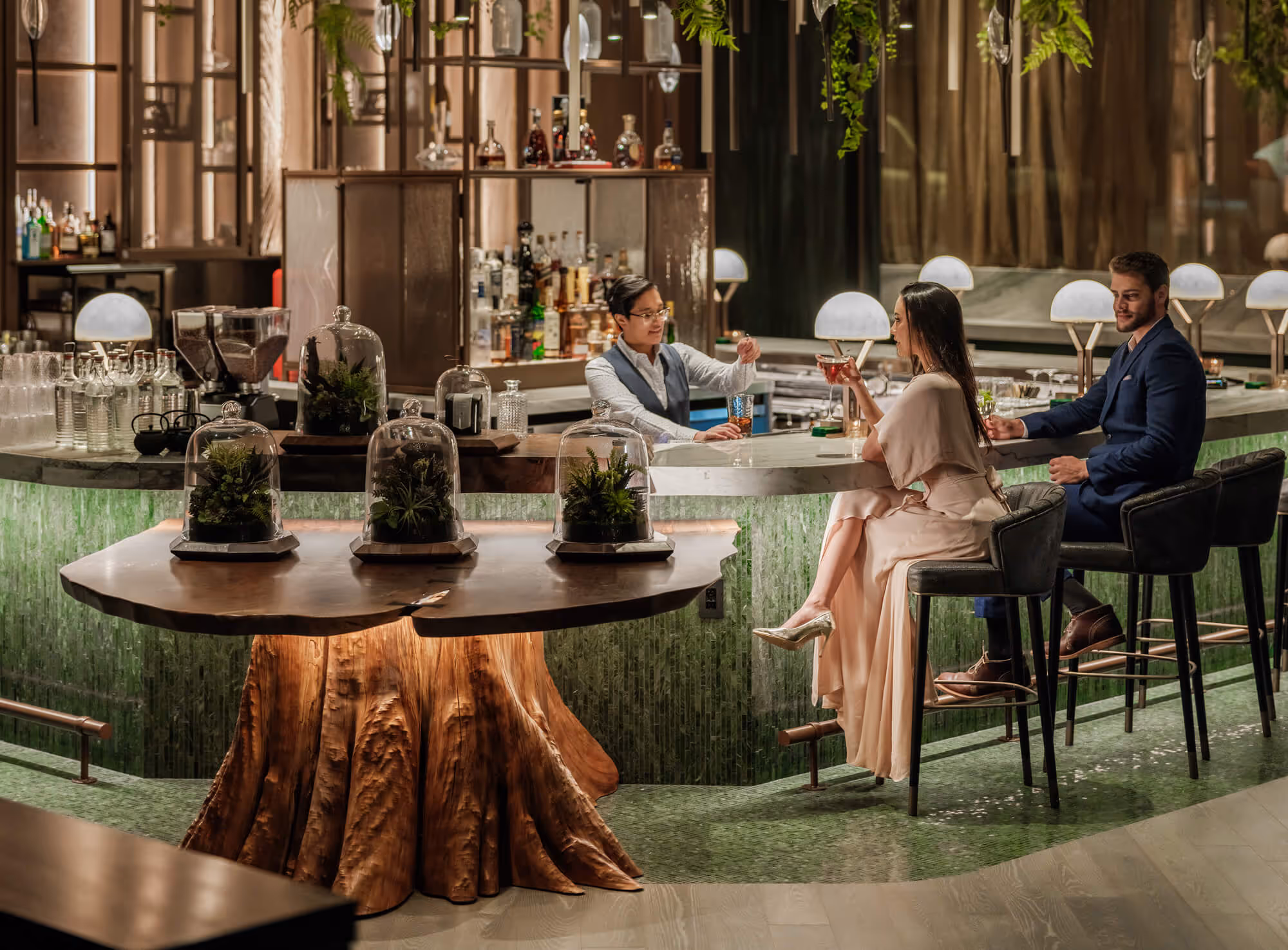 Elegant bar scene with a bartender serving drinks to a woman in a pink dress and a man in a blue suit sitting on high stools at Meadowrue