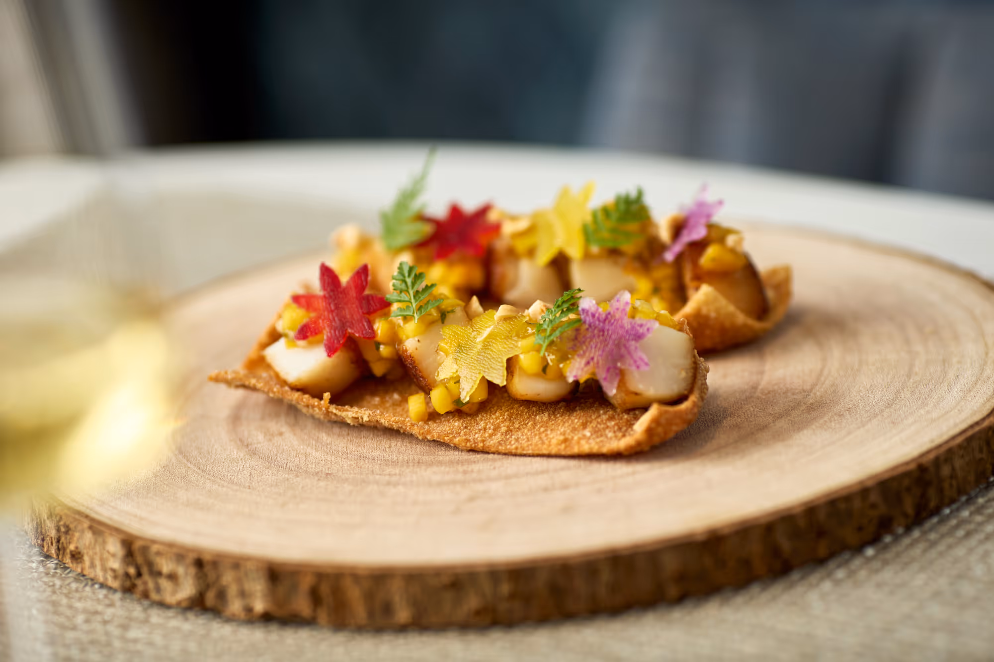 Gourmet appetizer with seared scallops, diced mango, edible colorful flower petals, and green herbs on a crispy base served on a wooden platter.
