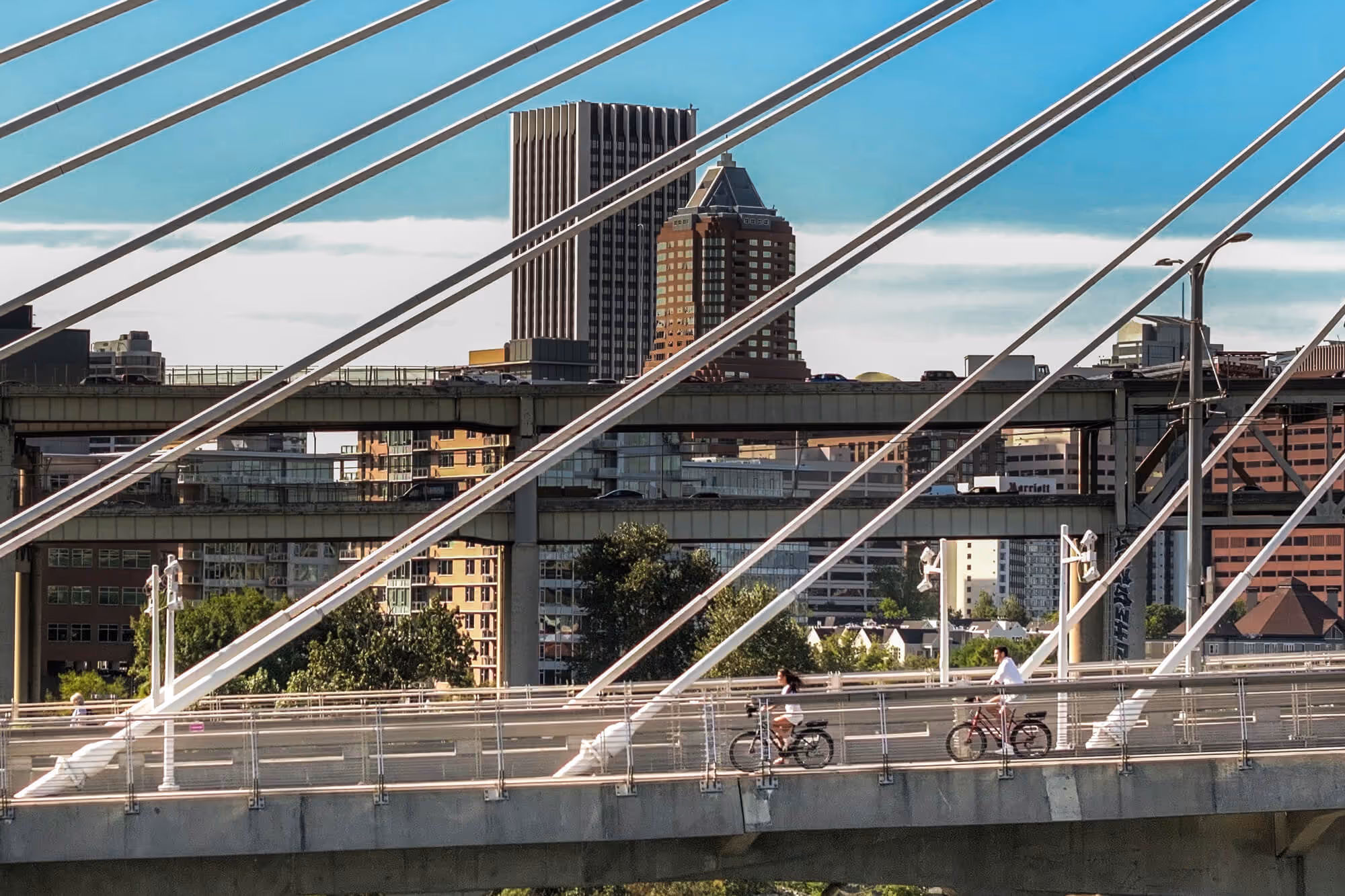 Two cyclists riding across the Tilikum Crossing Bridge with steel cables, city skyline and highway overpasses in the background under a blue sky.