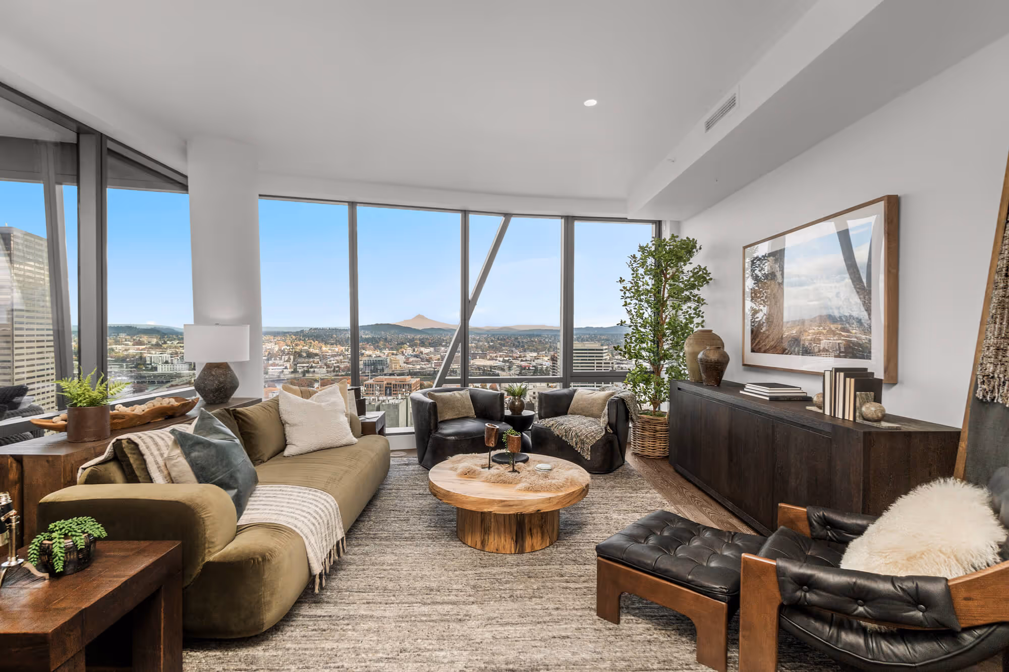 Modern living space in a Residence with floor-to-ceiling windows overlooking the Portland skyline and Mt. Hood, featuring a green couch, two black armchairs, a round wooden coffee table, and a wooden sideboard with plants and decor.