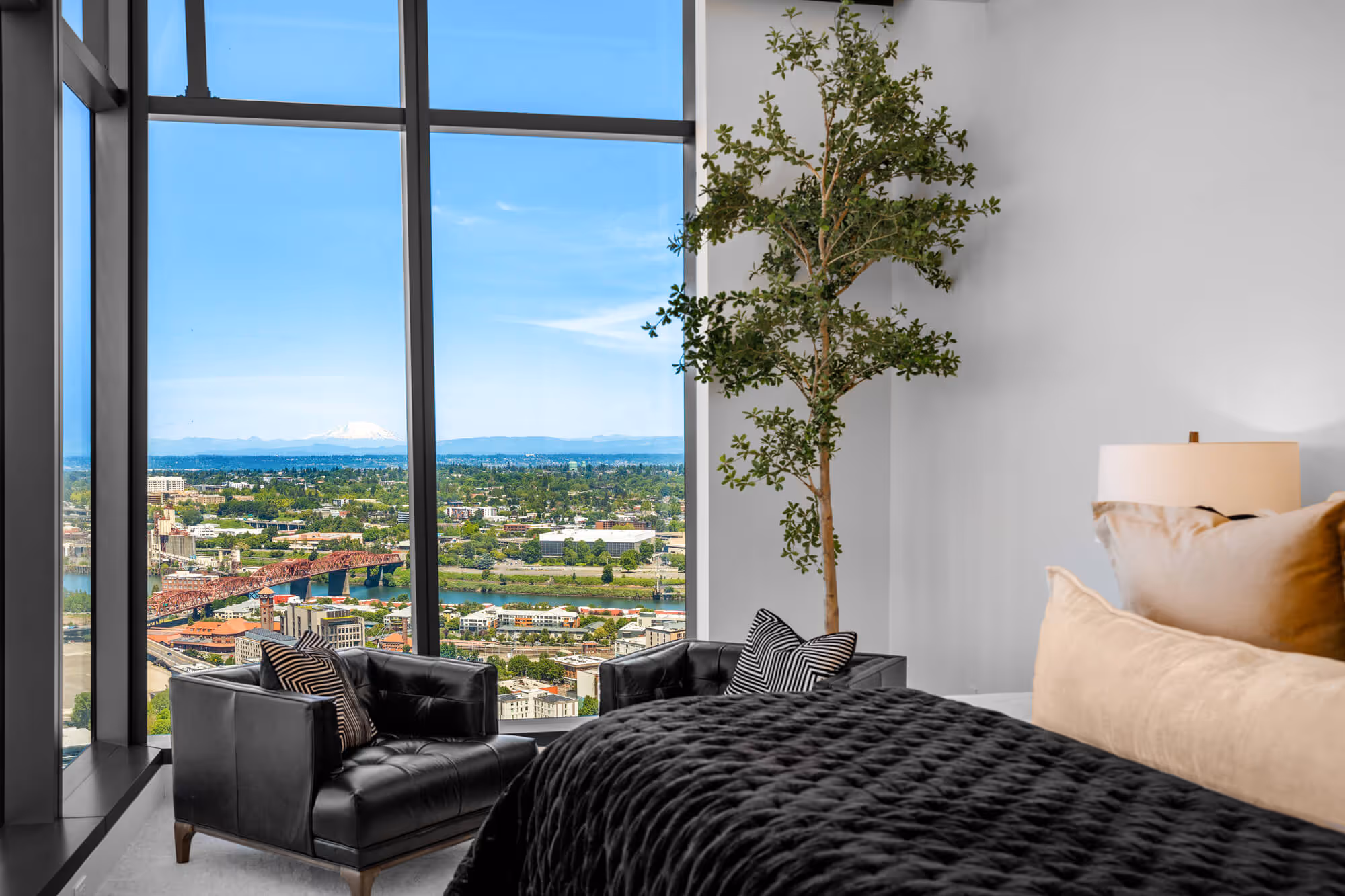 Penthouse bedroom with black leather chairs, large plant, and floor-to-ceiling windows showing the Portland skyline and distant Mount St Helens.