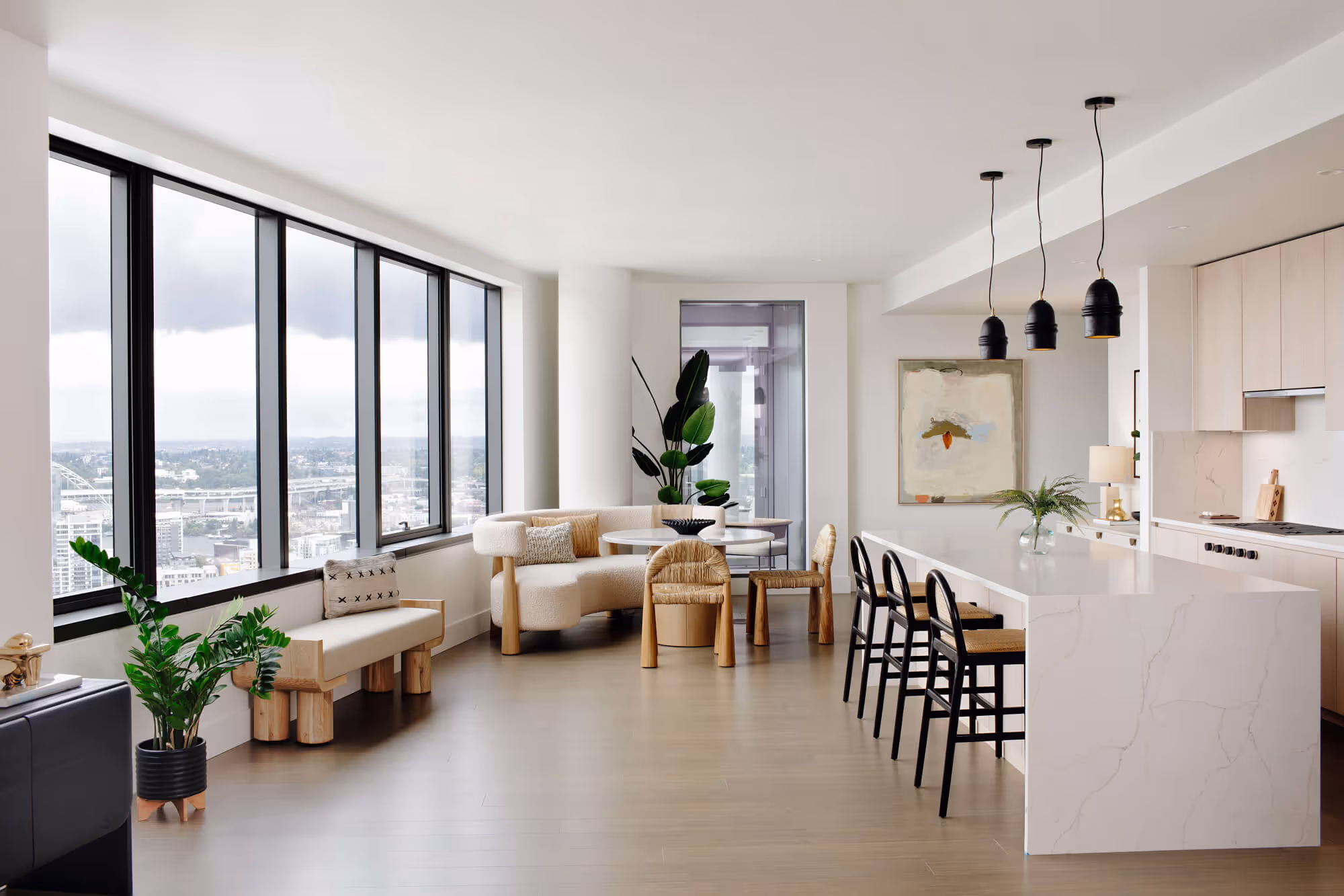 Bright modern kitchen and dining area in a Premium Residence with white marble island, black bar stools, curved beige sofa, wooden chairs, and large windows overlooking the Portland cityscape.