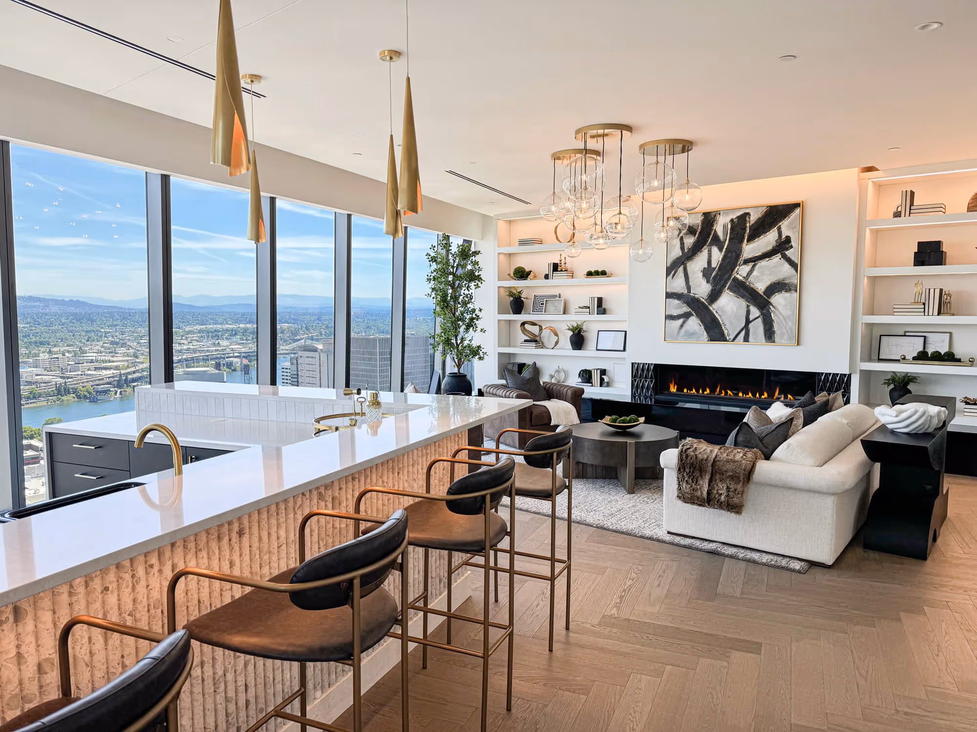 Open pPenthouse living area with large windows overlooking the Portland skyline, featuring a white sofa, fireplace, abstract painting, and bar stools at a marble island.