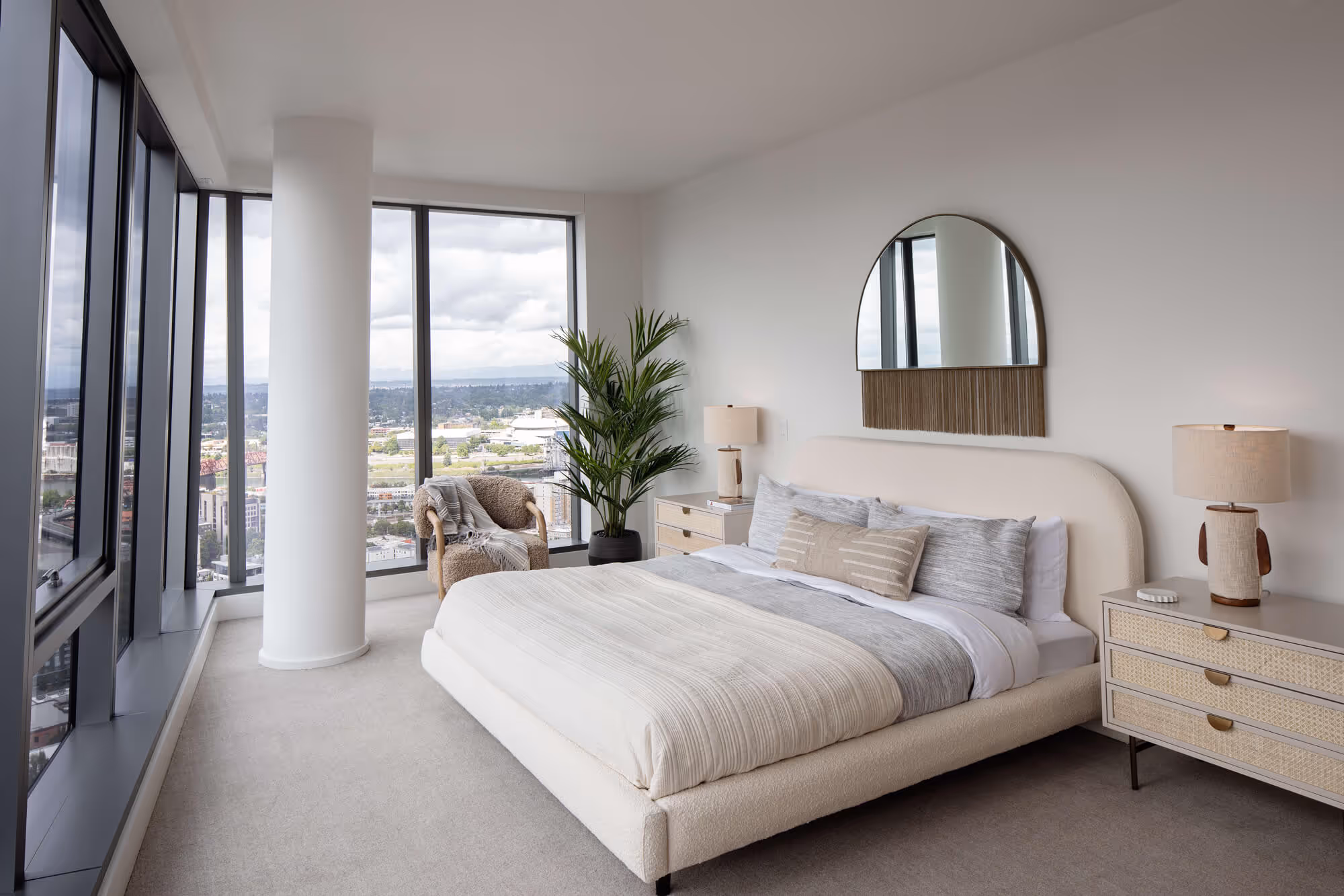 Premium Residence bedroom with large windows, a king-size bed with neutral bedding, two bedside tables with lamps, a round wall mirror, an armchair, and a tall indoor plant.