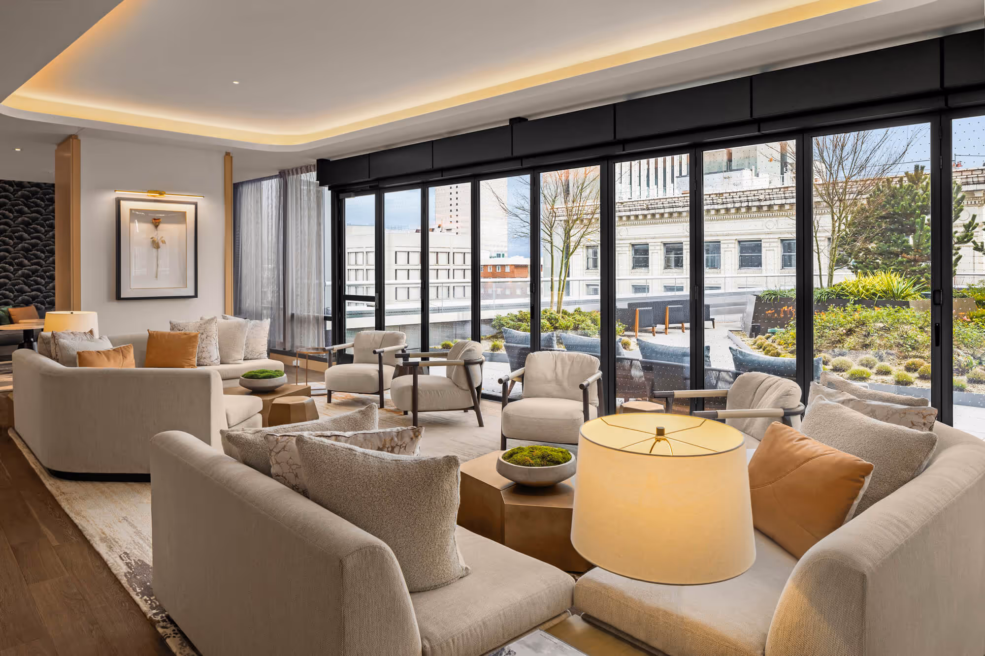 Seating area in Exclusive Owners' Lounge with beige sofas, armchairs, and window walls overlooking the rooftop terrace and surrounding buildings.