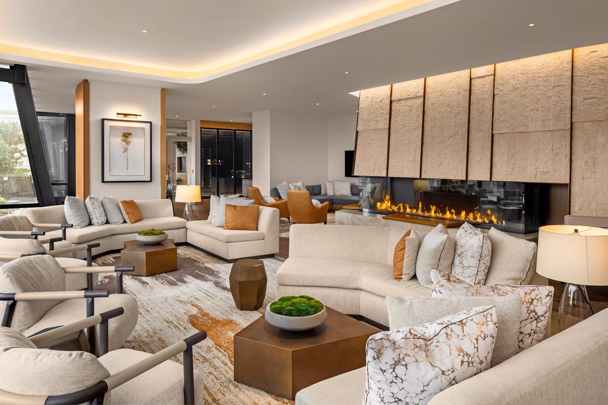 Exclusive Owners' Lounge with beige sofas and armchairs, decorative pillows, wooden coffee tables, a long fireplace, and warm lighting.
