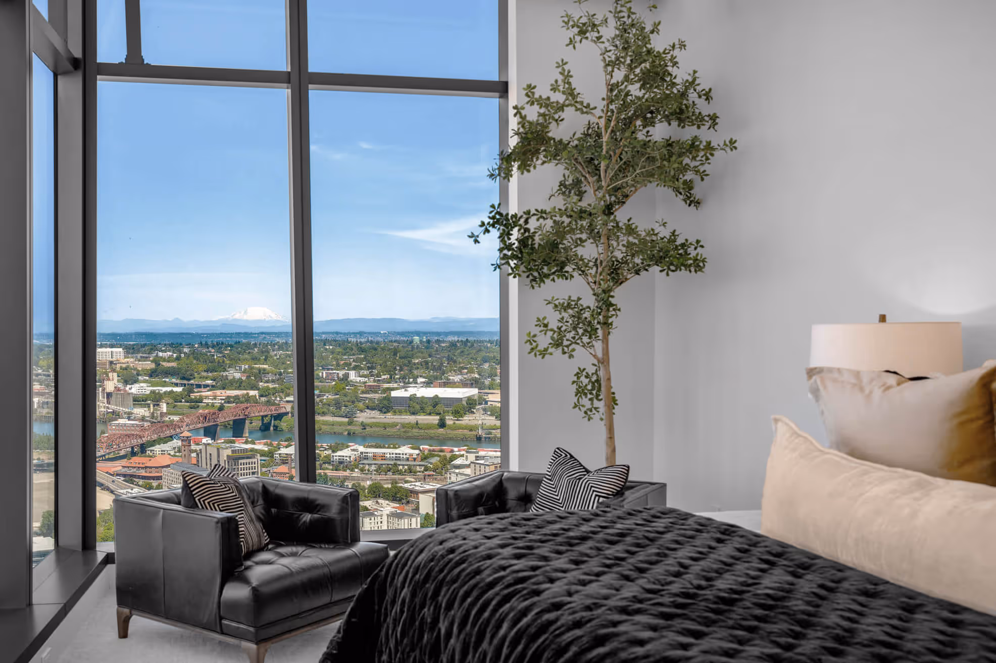 Penthouse bedroom with black leather chairs, large plant, and floor-to-ceiling windows showing the Portland skyline and distant Mount St Helens.