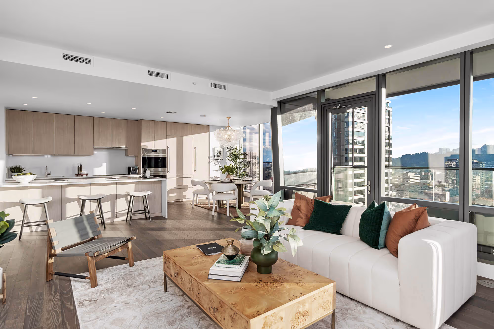 Open living space in a Residence with a white sofa adorned with green and orange pillows, wooden coffee table with books and a vase, and a kitchen with light wood cabinets and bar stools below large windows showing the Portland cityscape view.