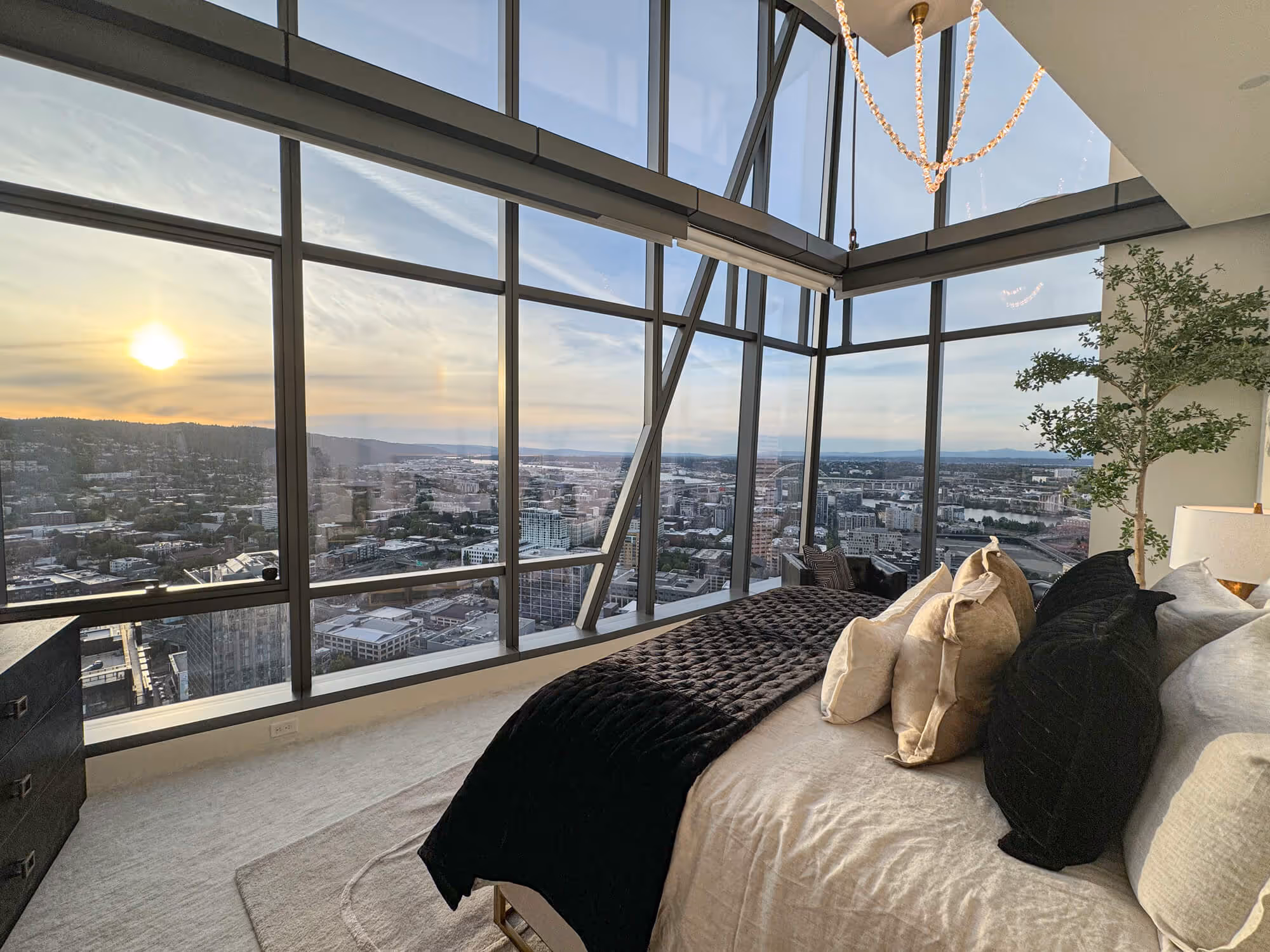 Penthouse bedroom with large floor-to-ceiling windows showing the Portland skyline at sunset, a bed with black and beige pillows, and a chandelier overhead.